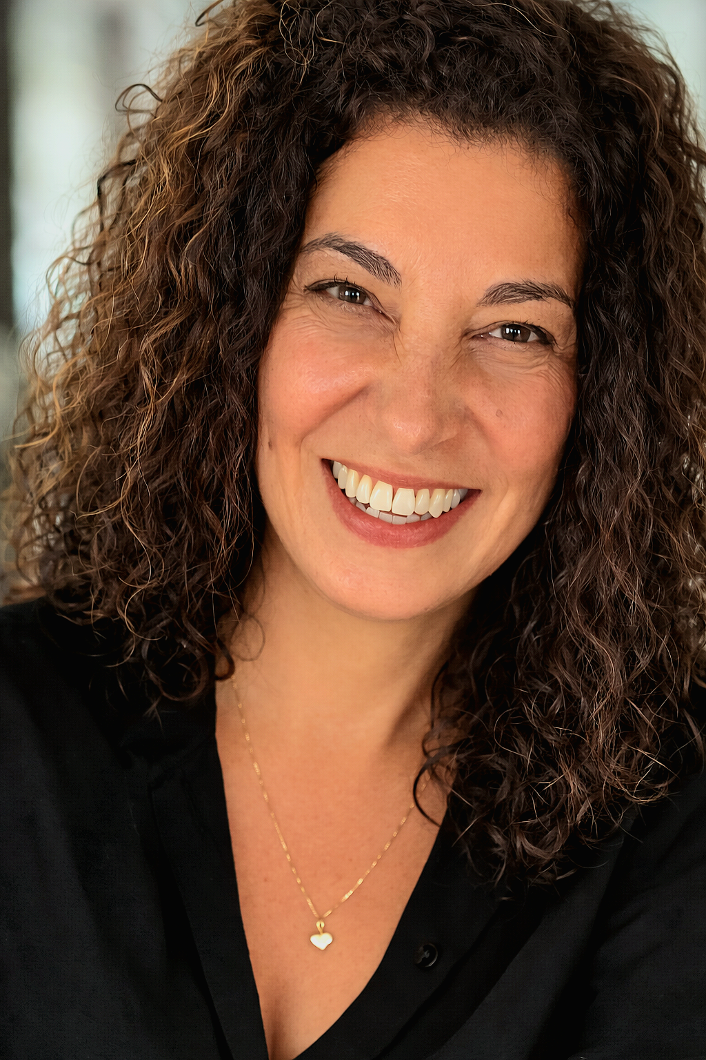 A woman with curly brown hair smiling at the camera, wearing a black top and a gold necklace with a heart-shaped pendant.