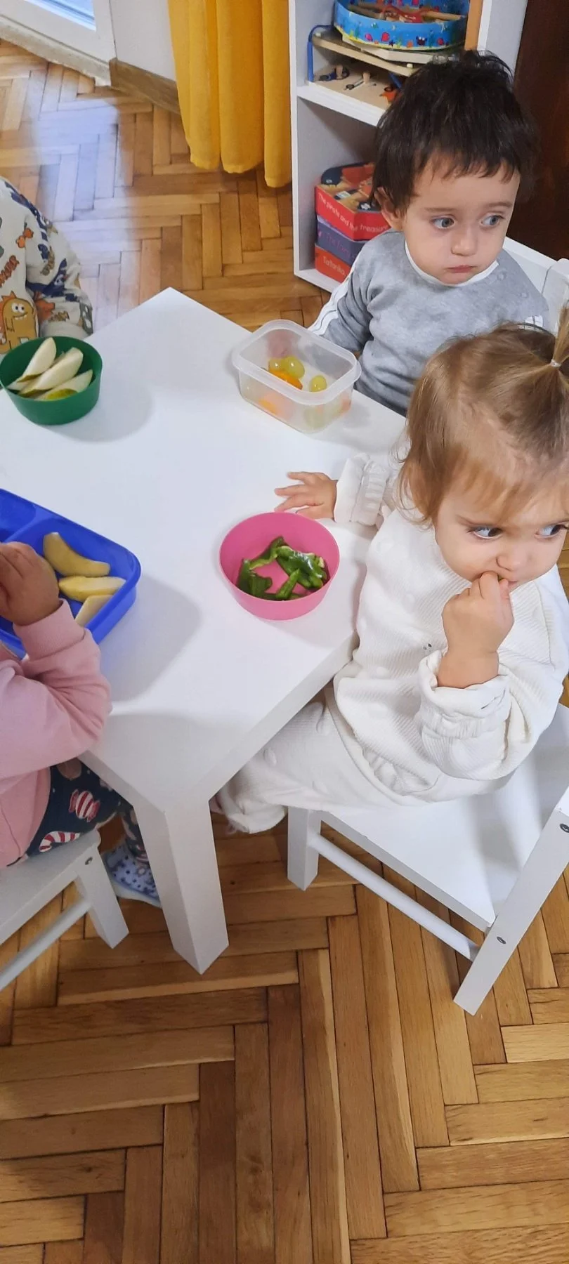 Children sitting at a white table with bowls of fruits and vegetables, including green peppers, bananas, and grapes, in a room with wooden parquet floor and a white bookshelf in the background.