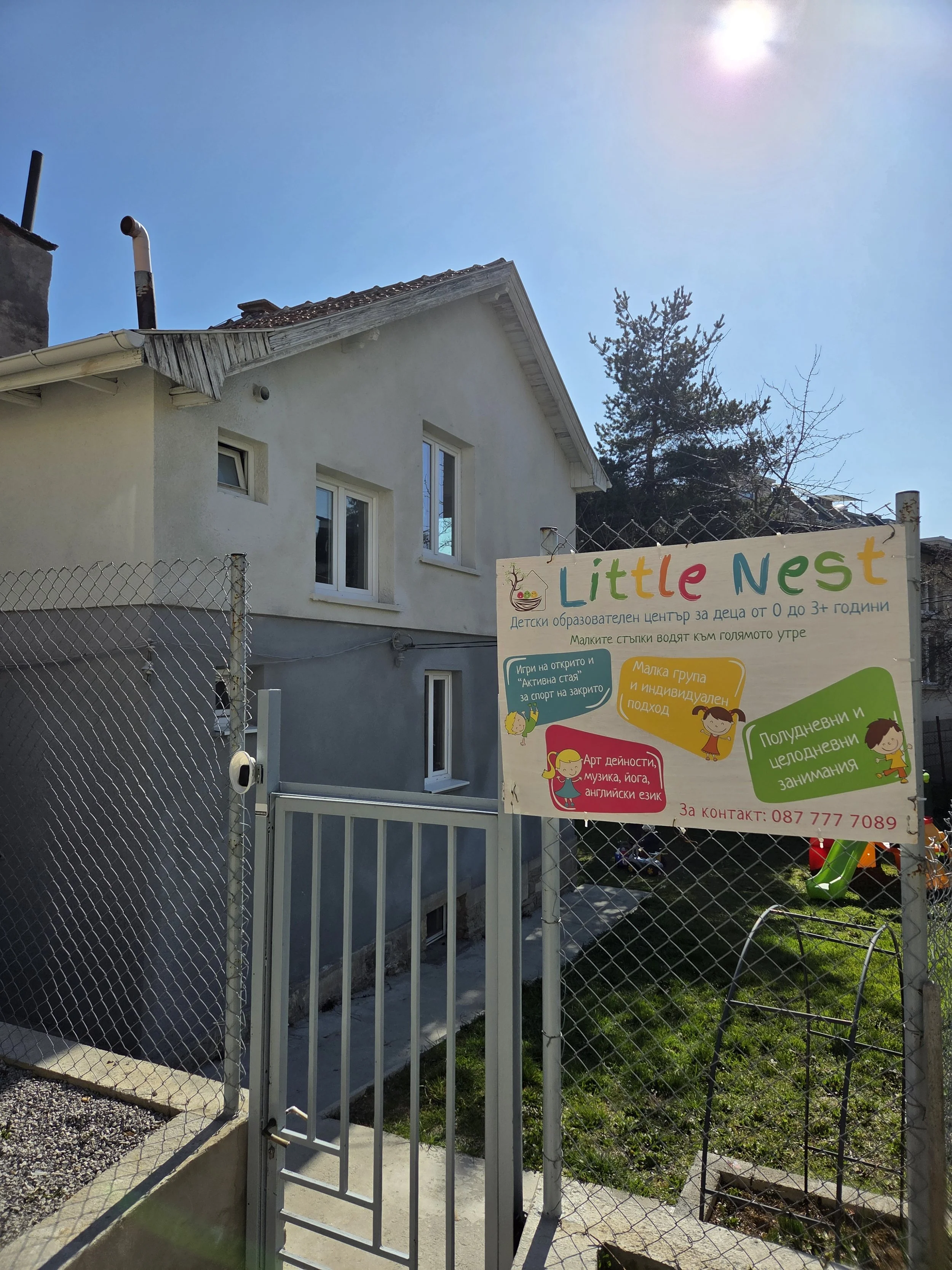 The image shows the exterior of a house with a sign on a fence that reads "Little Nest," a children's educational center. The sign has colorful text and cartoon illustrations of children, indicating activities like arts, music, yoga, and English lang