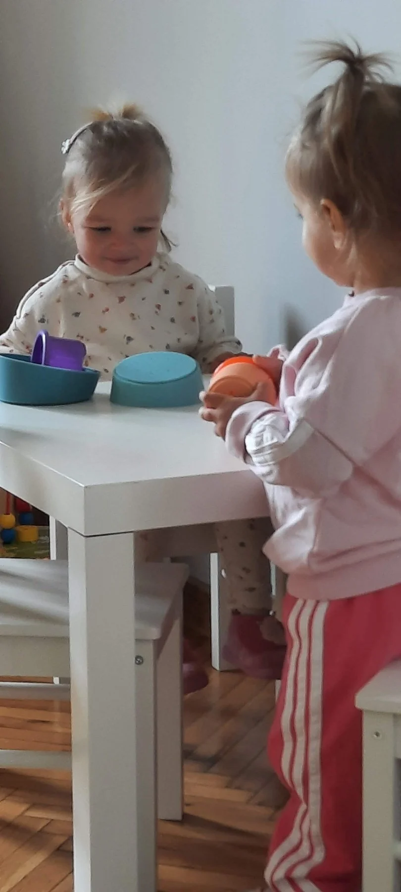 Two young girls with light-colored hair playing with plastic containers and lids at a white table in a room with wooden flooring.