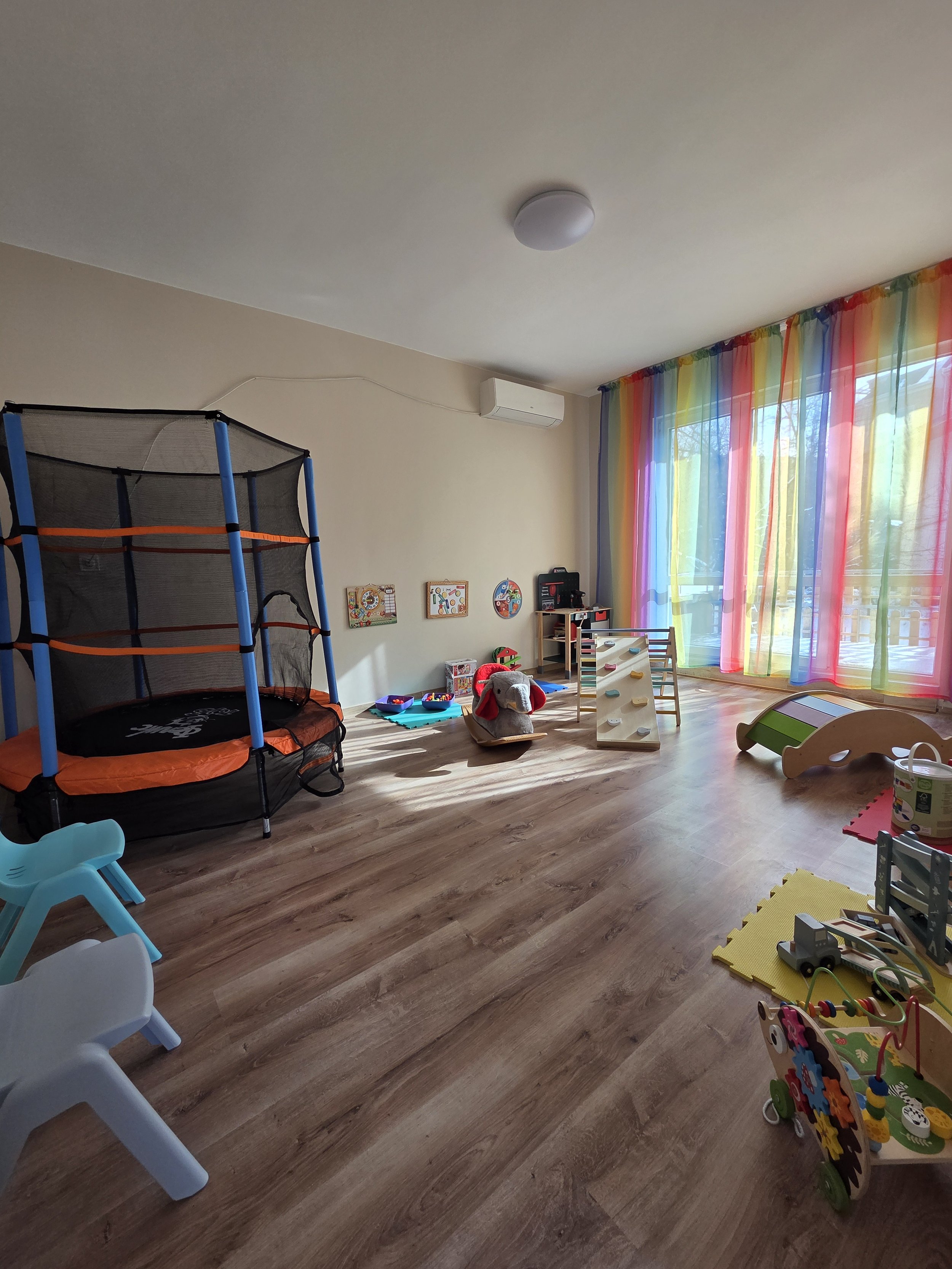 Bright colorful children's playroom with rainbow curtains, trampoline, rocking horse, toys, and small chairs.