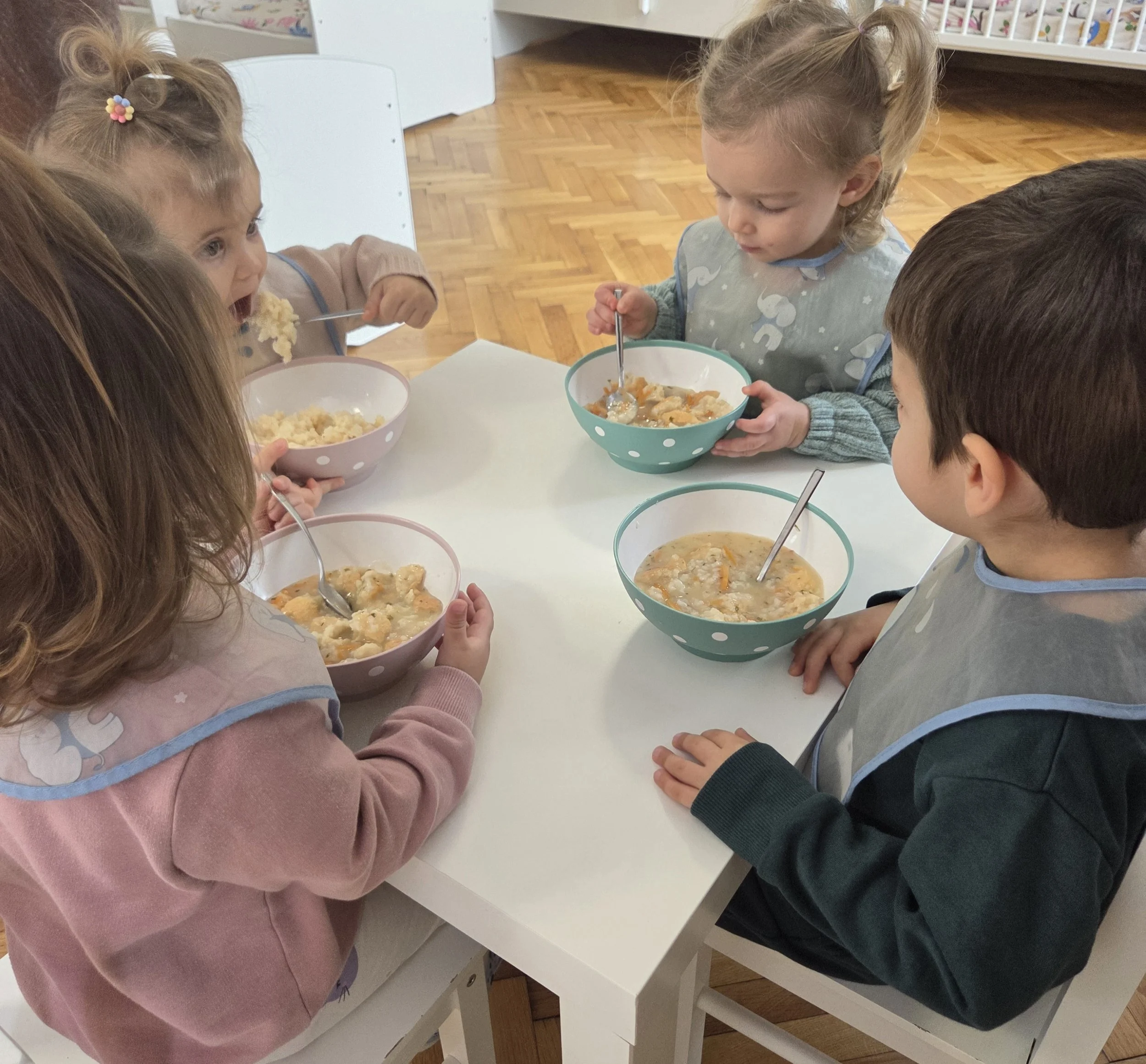 Four children sitting at a white table eating from bowls of soup or stew. The children are wearing bibs and are using spoons. The setting appears to be a classroom or daycare with a wooden floor.