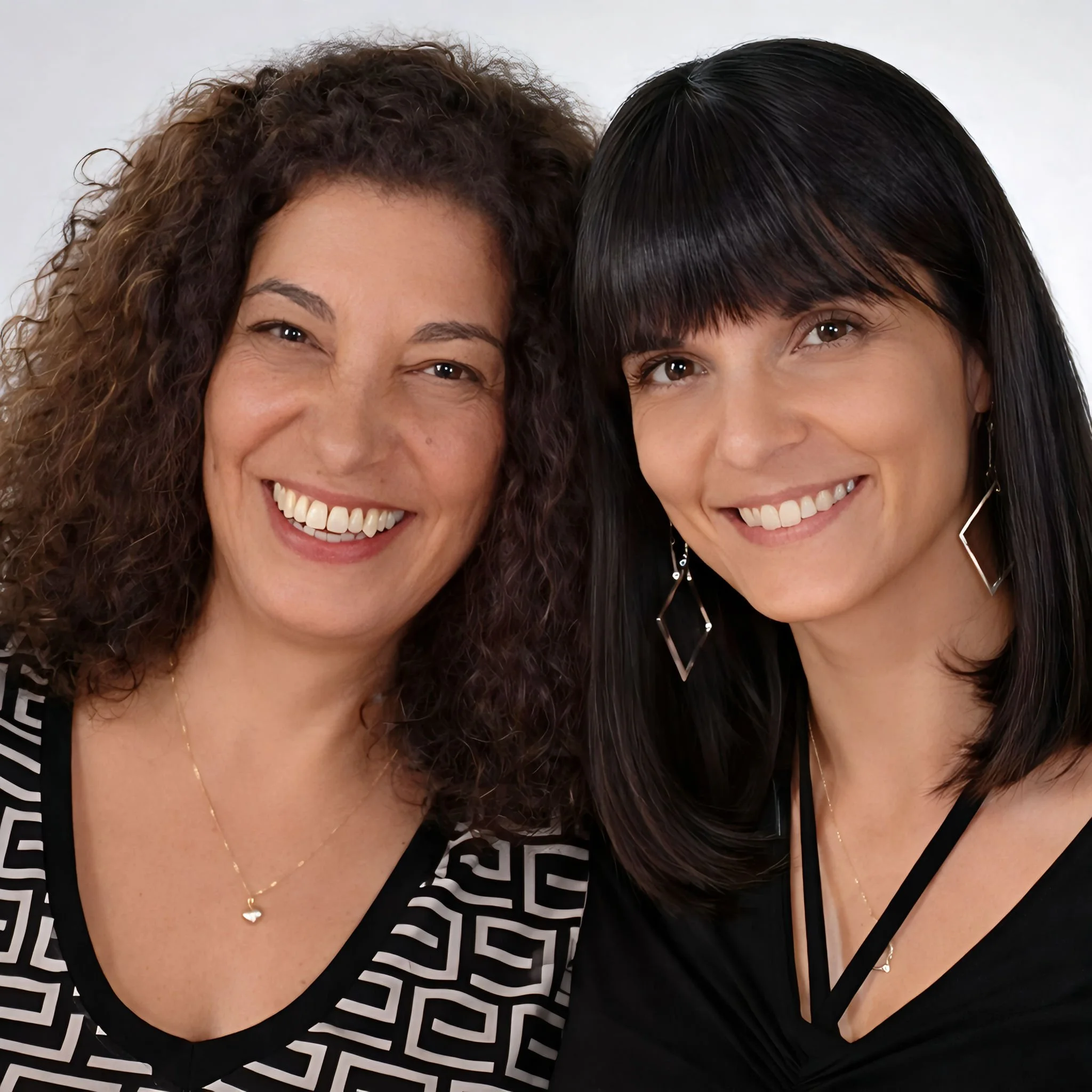 Two women smiling and posing closely together, one with curly hair and the other with straight dark brown hair and bangs.