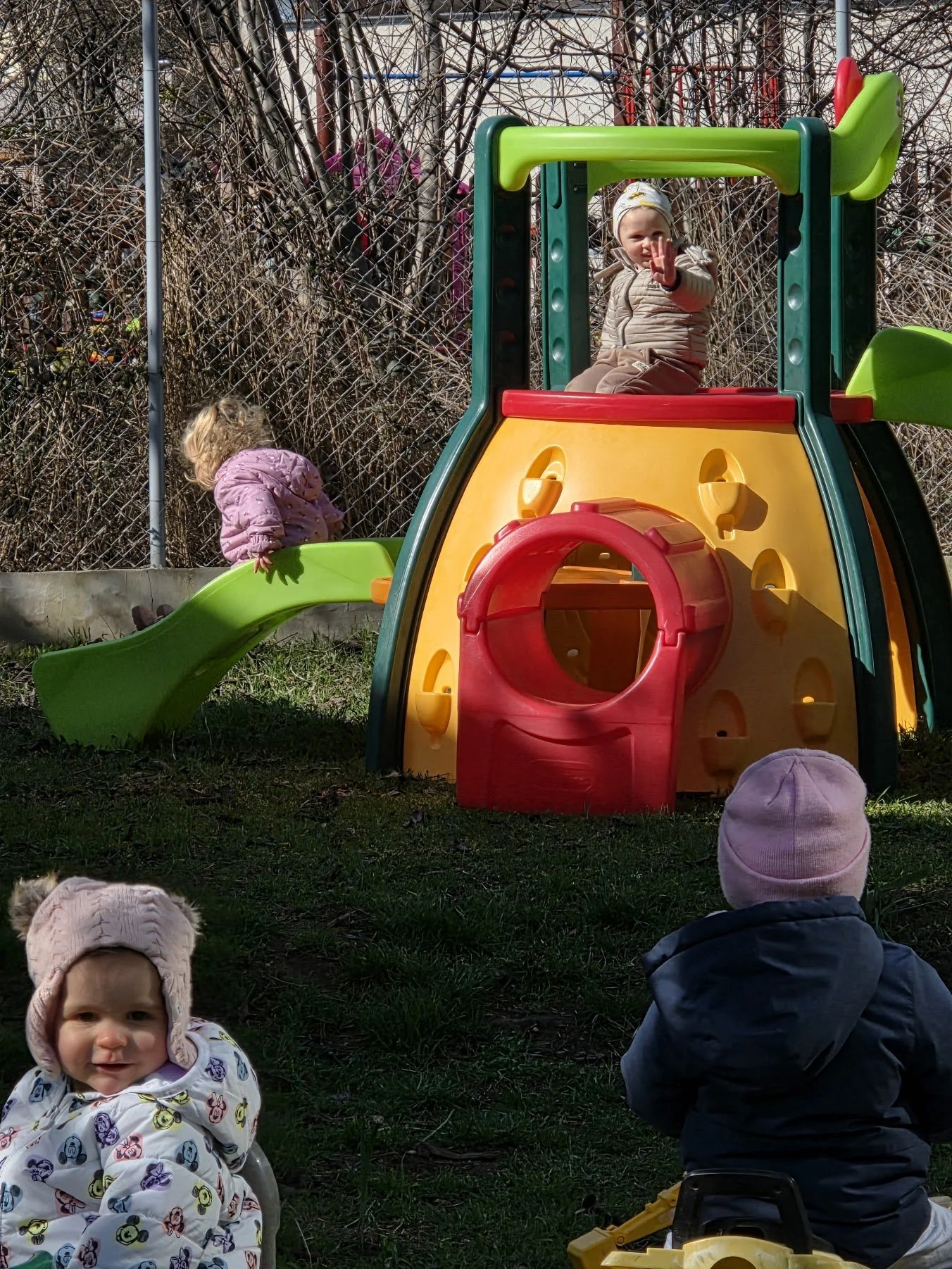 Four children in jackets playing at a playground with a colorful plastic slide, one child sitting at the top waving.
