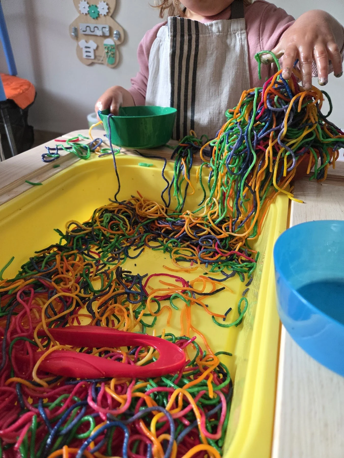 Child playing with colorful rubber bands in a sensory bin, wearing an apron, with a green and a blue bowl nearby.