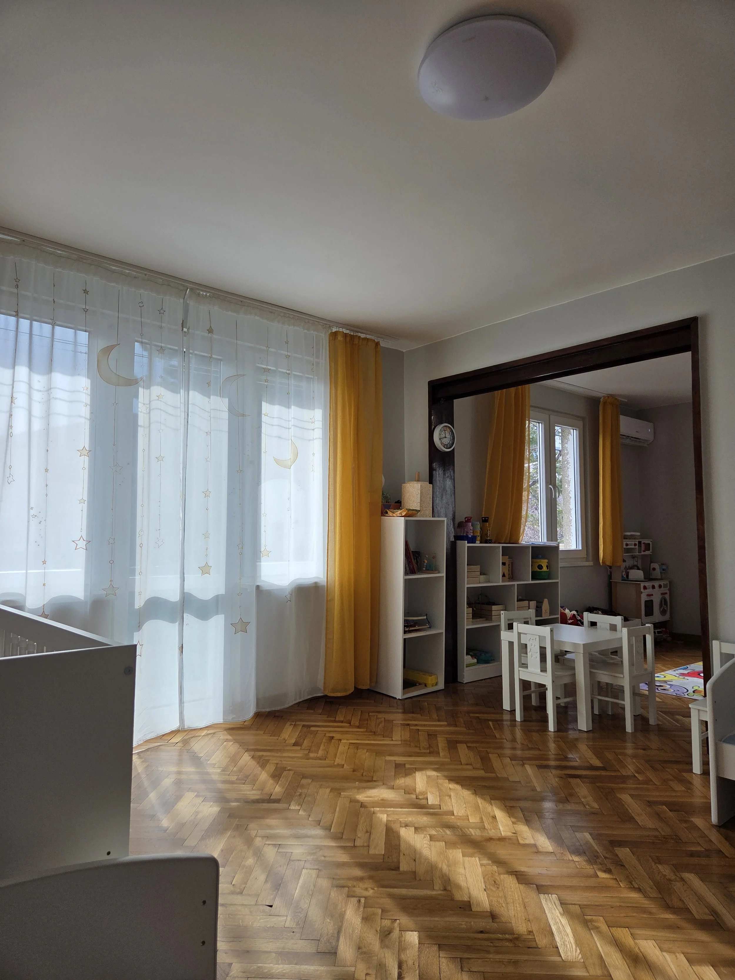 A room with large windows covered with sheer curtains with moon and star designs and yellow curtains, wooden parquet flooring, and children’s furniture including small white tables and chairs, white storage shelves with toys and books, and a play are