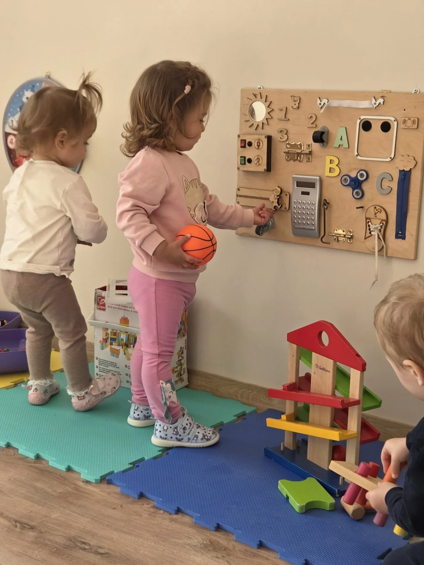 Children playing with educational toys in a playroom, including a wooden activity board and a colorful ramp structure.