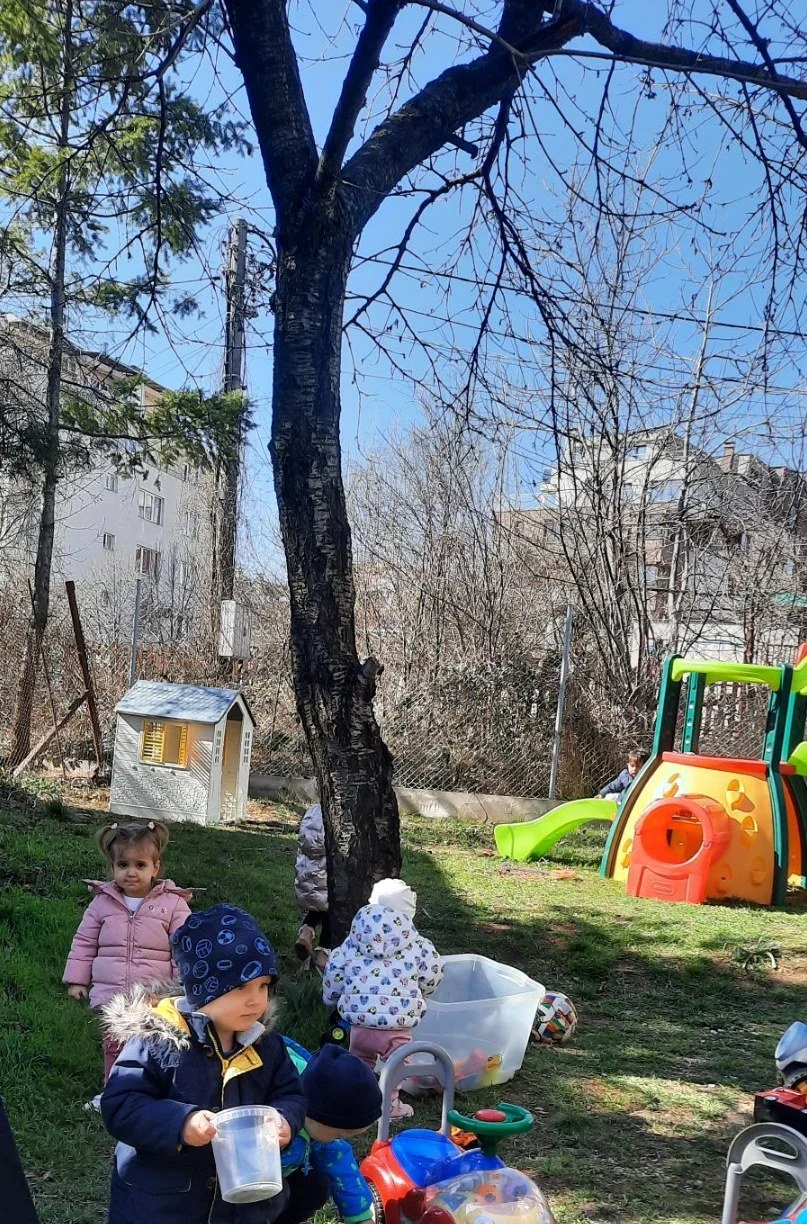 Children playing outdoors on a grassy area with a toy house, slide, and various toys under a large tree.