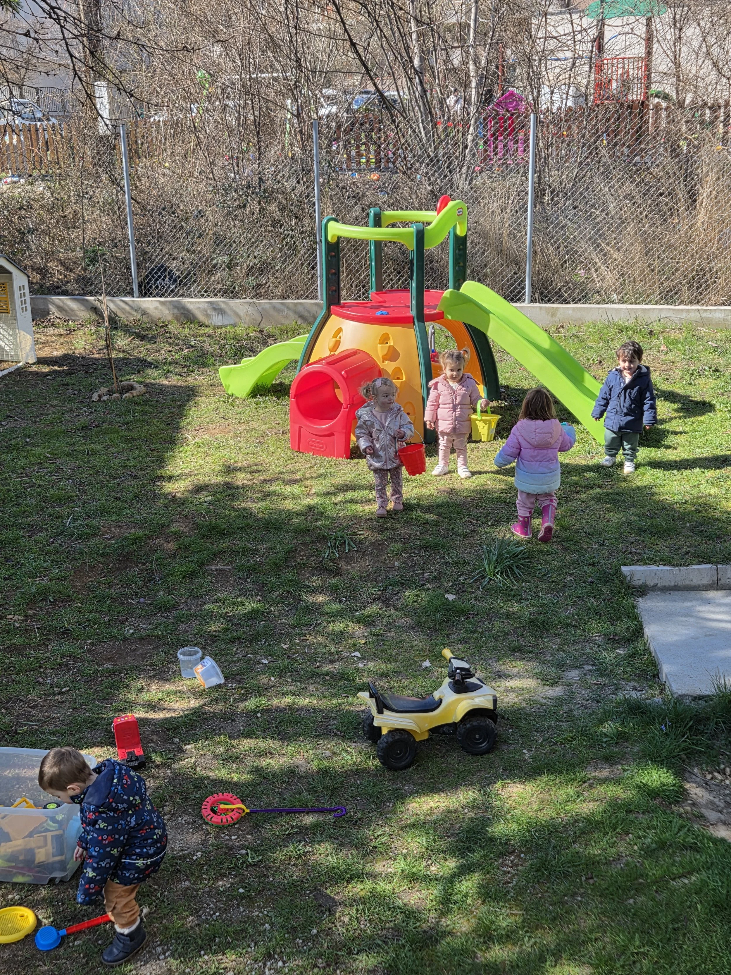 Children playing on a colorful plastic playground with slides and tunnels, holding toy buckets, in a grassy yard enclosed by a chain-link fence.