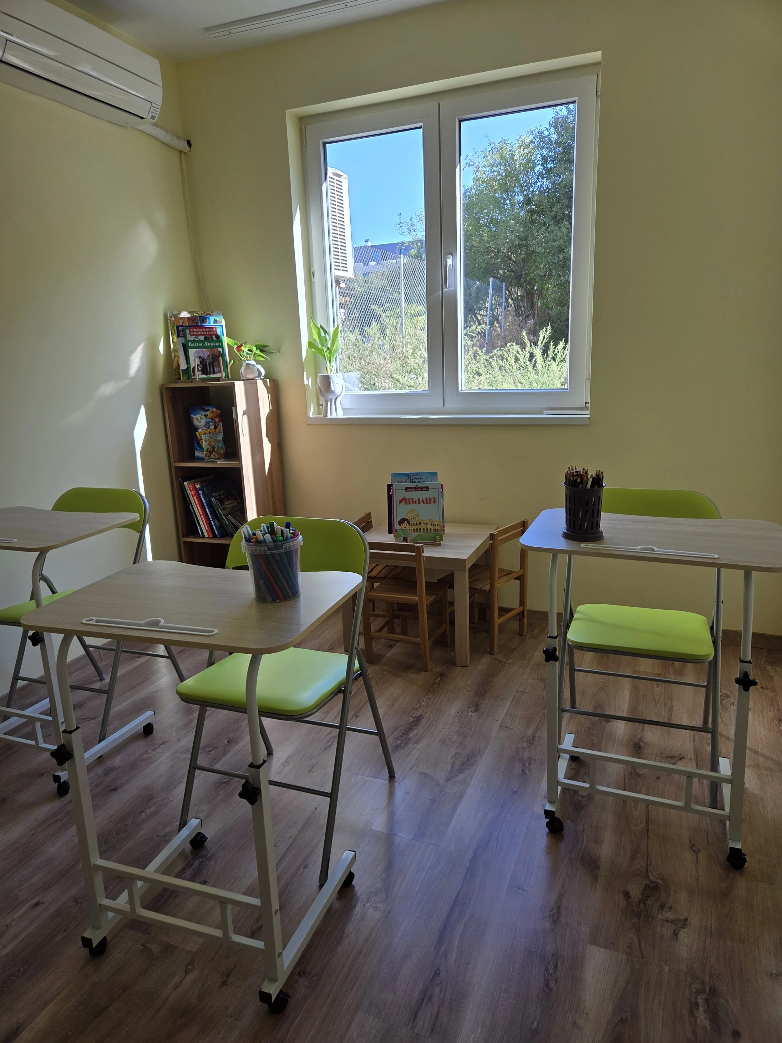 A bright classroom with light green chairs, small tables with containers of colored markers, a window showing greenery outside, and a wooden bookshelf with books and plants.