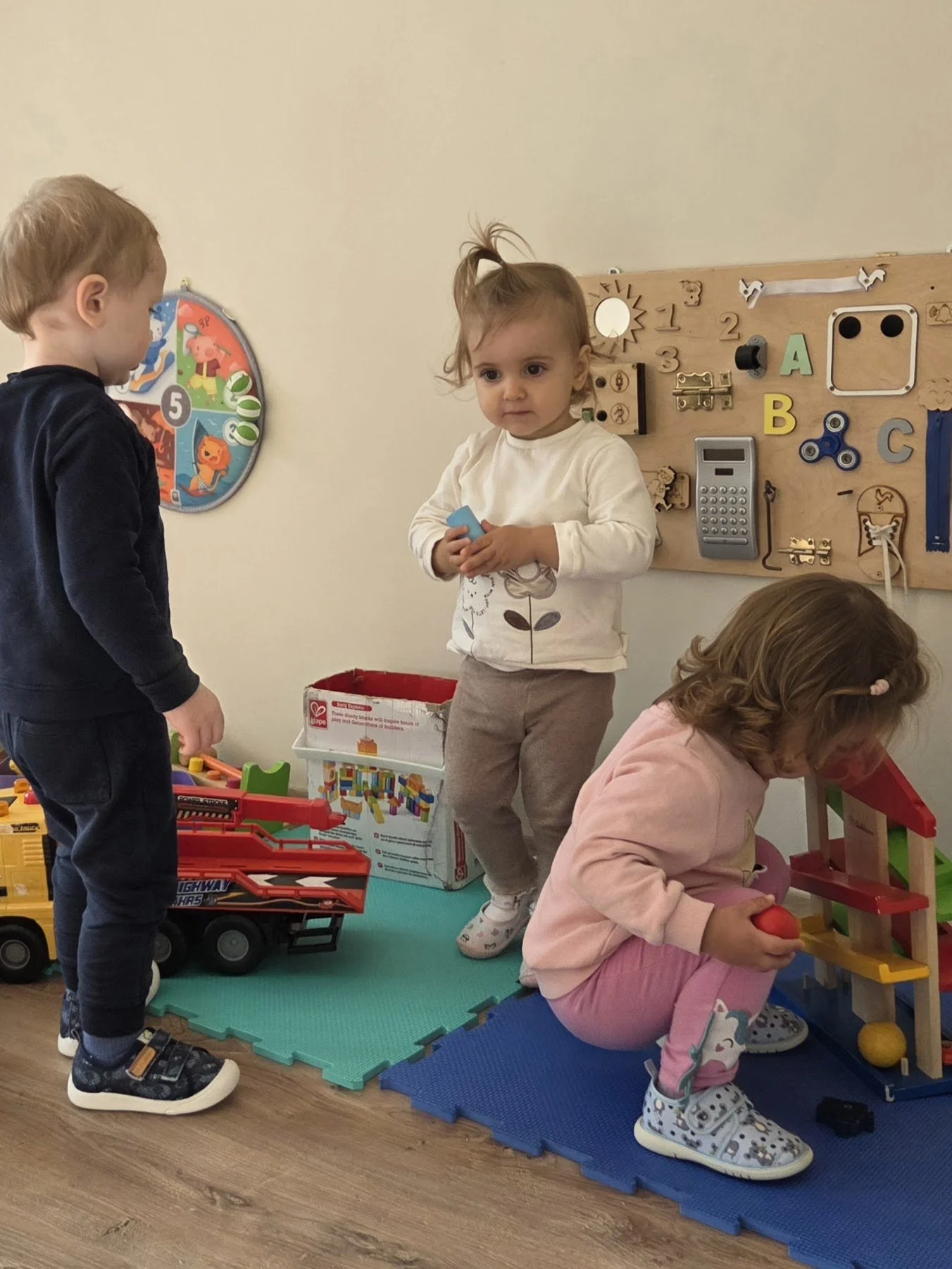 Three young children playing with toys in a playroom. One girl in pink pants is seated, holding a red object, while a girl in beige pants stands nearby, and a boy in dark clothes stands on the opposite side.
