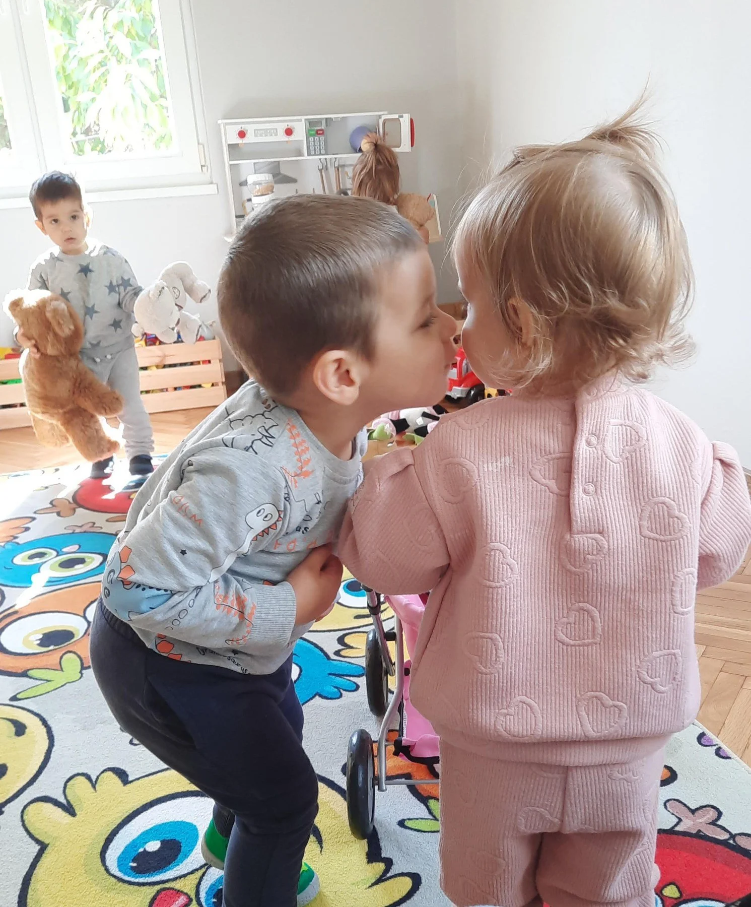 Two young children, a boy and a girl, sharing a kiss in a playroom. The boy is leaning forward, and the girl is facing him. In the background, a third child is playing with teddy bears, and there are toys and colorful patterns on the floor.