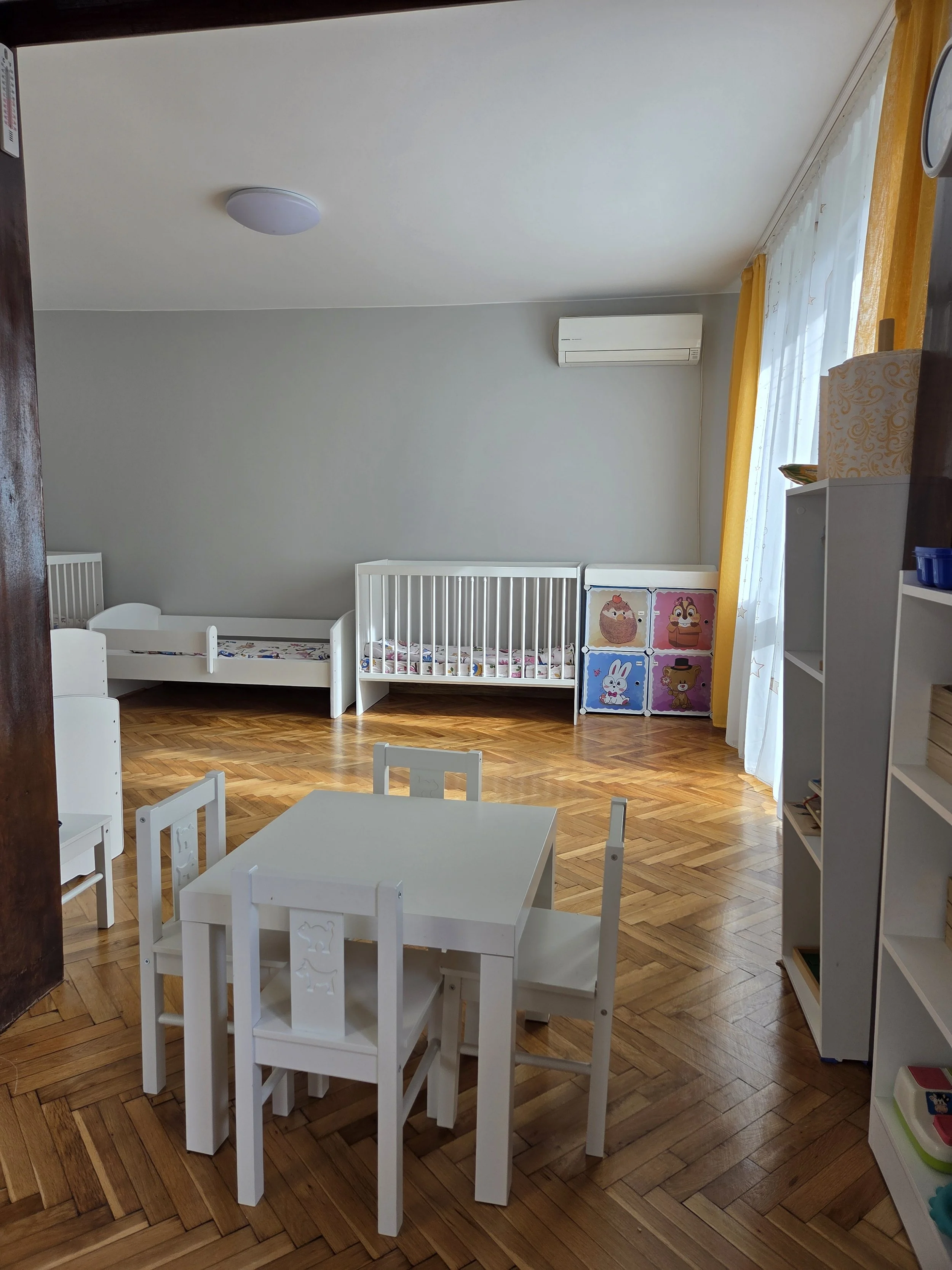 Children's room with two white cribs, a small white table with four chairs, a white shelf with children's books, a colorful storage unit with animal illustrations, and a window with yellow curtains.