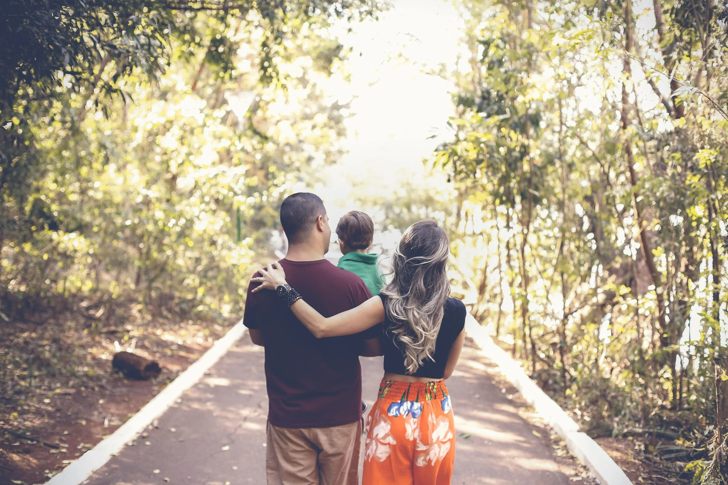 A family of three walking on a forest path, with a man, woman, and a child seen from behind, surrounded by trees and bright sunlight.