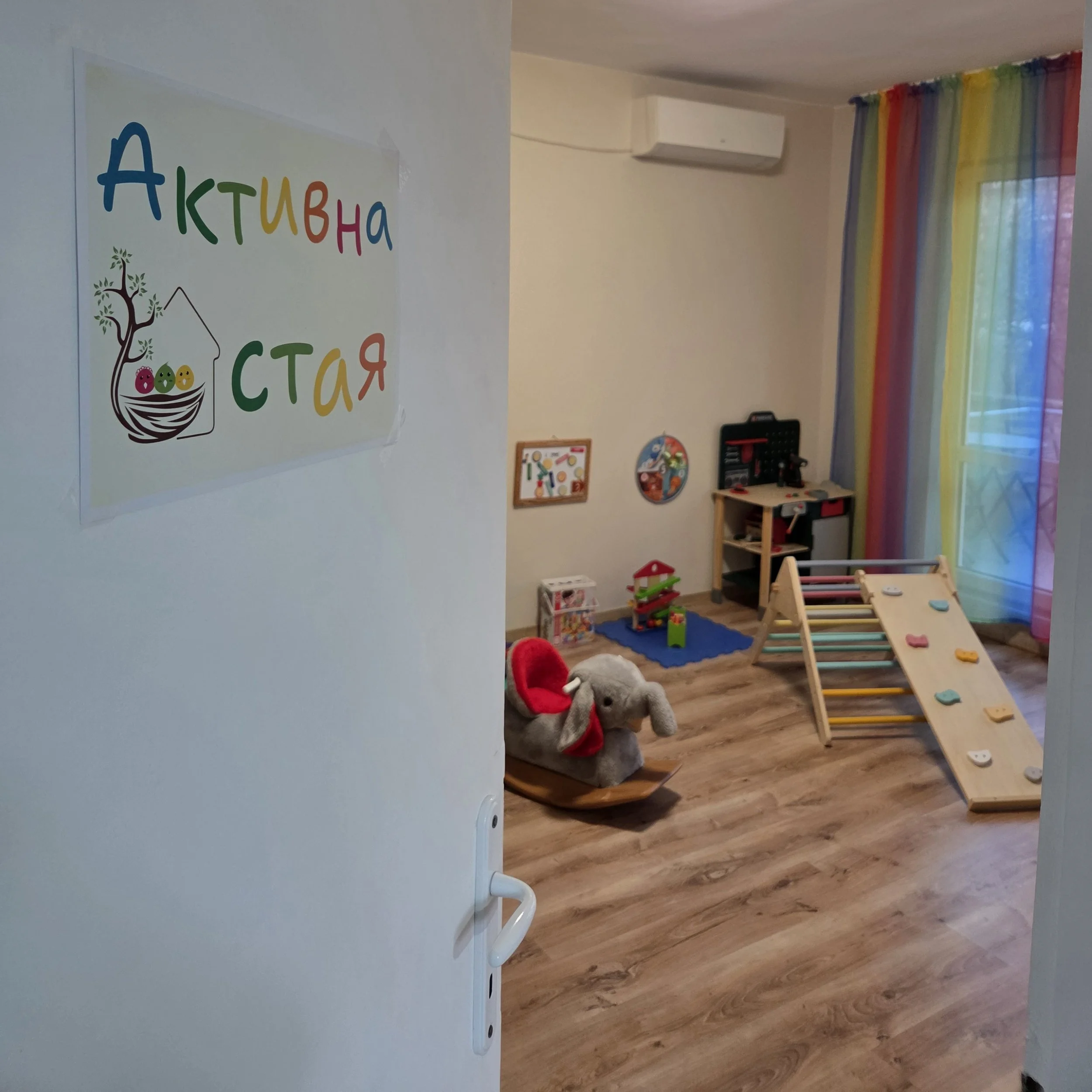 Children's activity room with climbing wall, stuffed elephant on a rocking toy, rainbow curtains, colorful toys, and a sign in Russian on the door that says 'Active Room'.