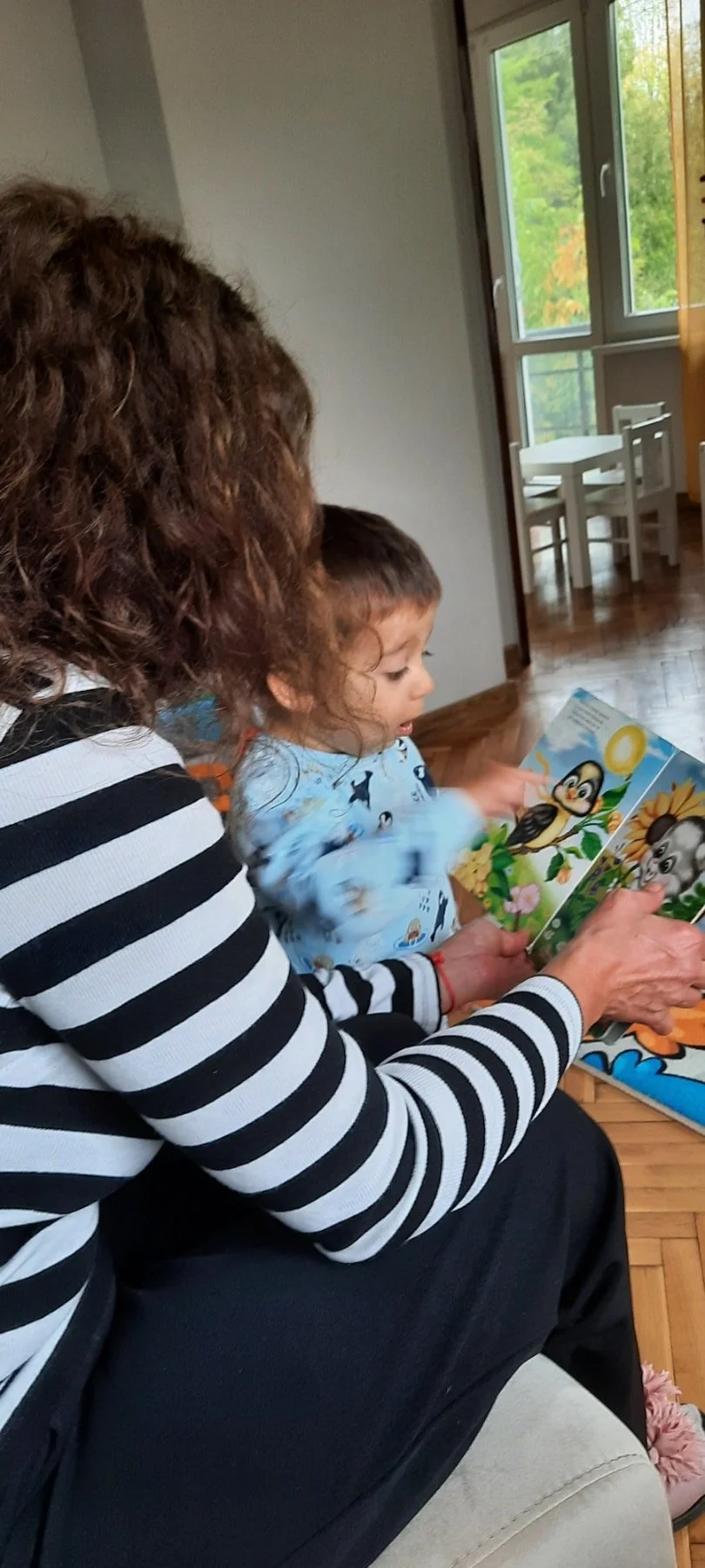 A woman with curly brown hair and a child with short brown hair are sitting together, reading a children's picture book about animals. The woman is wearing a black and white striped shirt, and the child is dressed in a light blue pajama top featuring