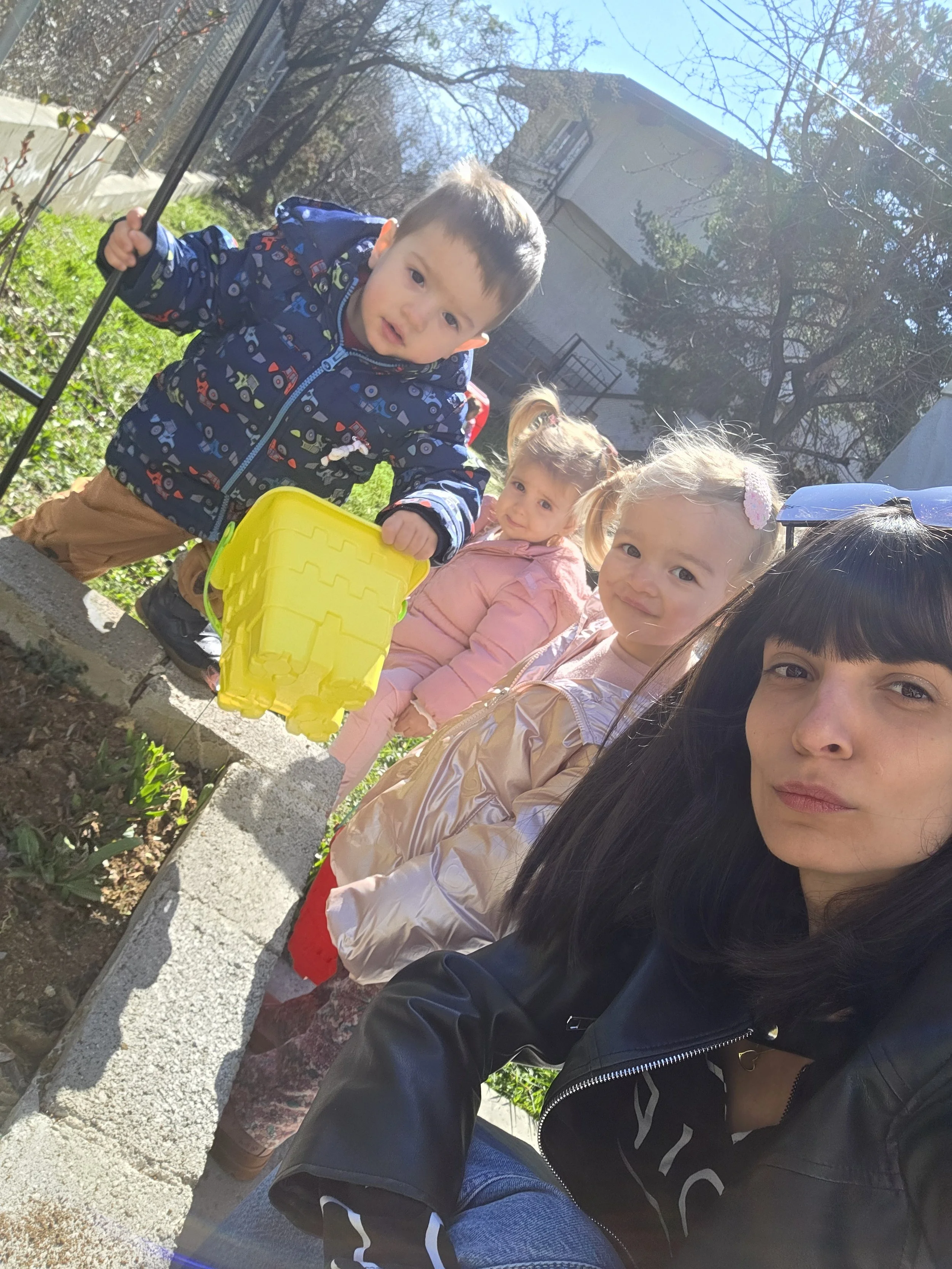 A woman taking a selfie with three young children outdoors on a sunny day, with a garden and house in the background. The children are dressed in jackets and are sitting or standing near a garden bed.