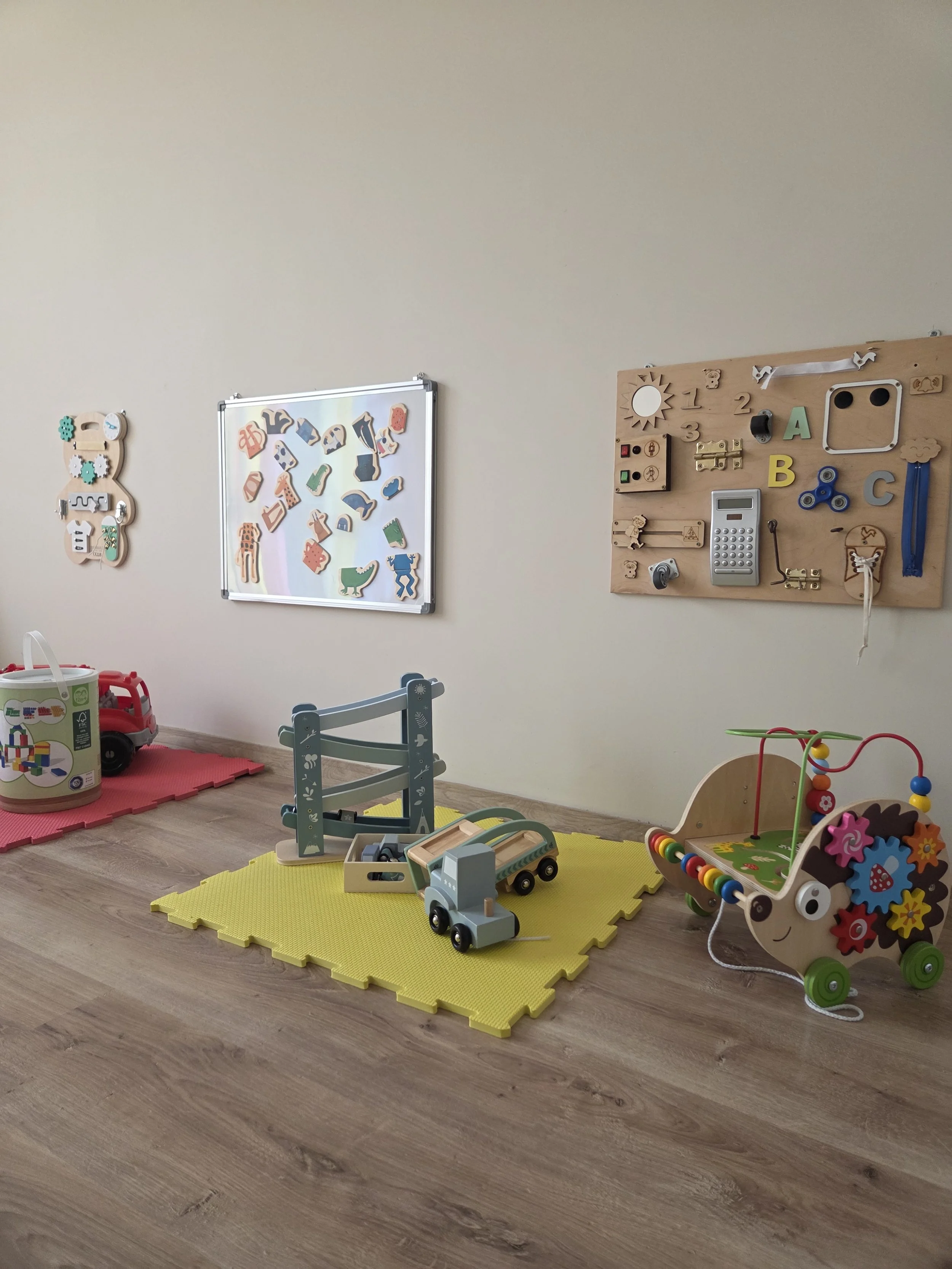 A children's playroom with wooden toys, a yellow foam mat, and colorful wall decorations, including a whiteboard with puzzles and a pegboard with educational toys.
