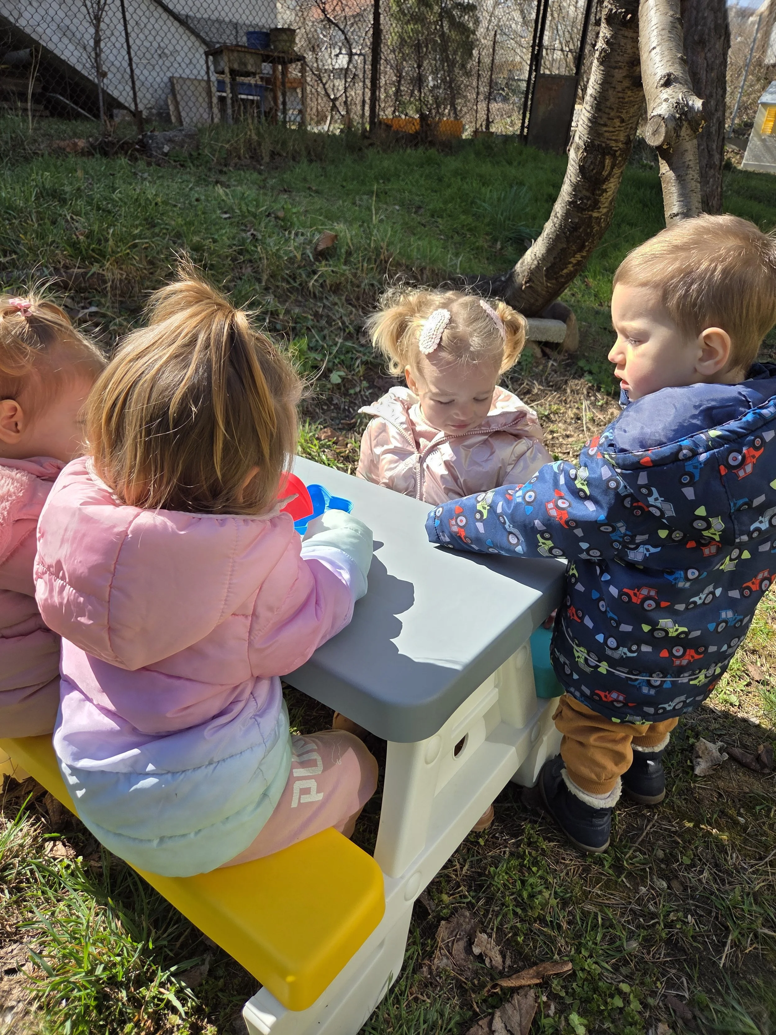 Four young children sitting and standing around a small plastic table outdoors on a sunny day, with three of the children sitting on a yellow bench and one standing. They are engaged in an activity, possibly playing with a toy or a container on the t