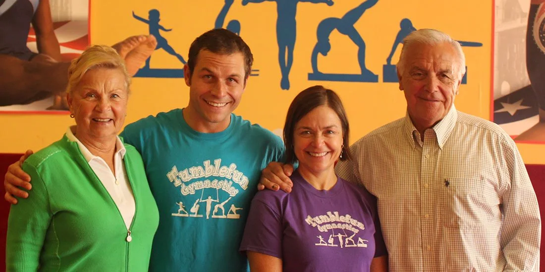 Four people smiling and posing for a photo in front of a yellow wall with silhouettes of gymnasts. Two women and two men, all wearing colorful casual shirts.