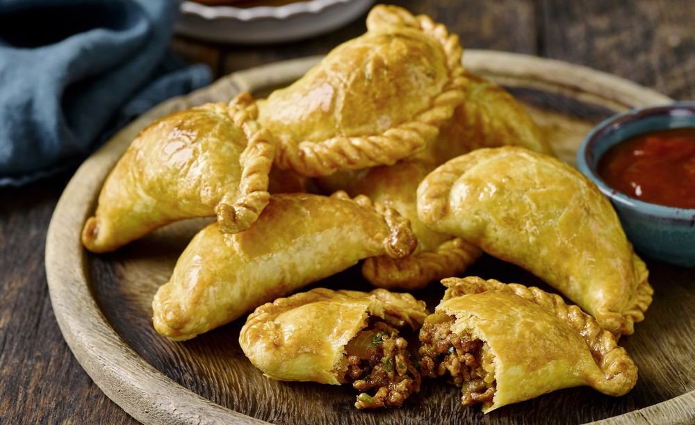 A wooden serving tray with cooked empanadas, one cut open to reveal ground beef filling, and a small bowl of red dipping sauce.