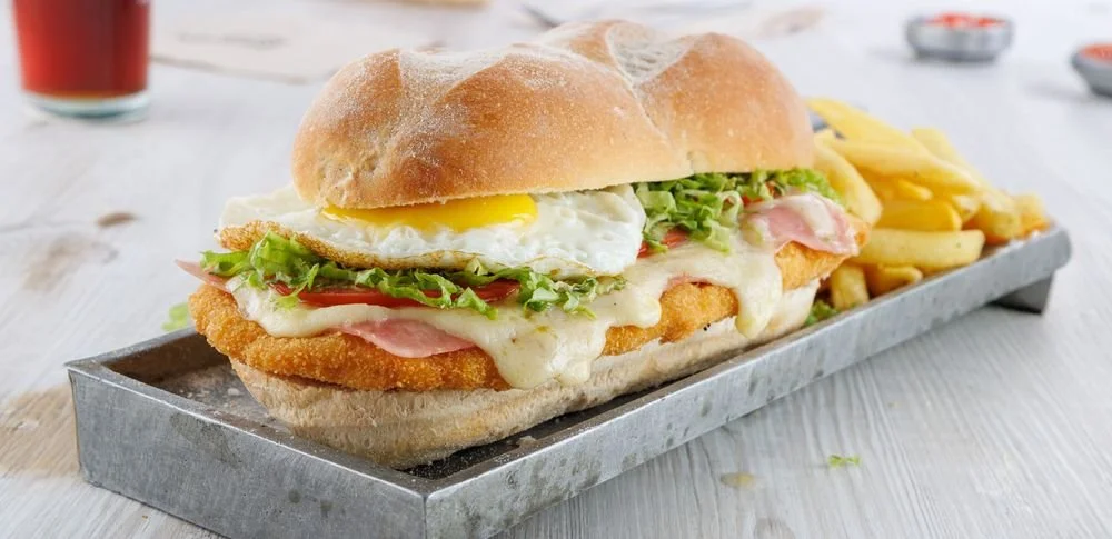 A breaded fish fillet sandwich with a fried egg, lettuce, tomato, cheese, and ham, served with French fries on a metal tray.