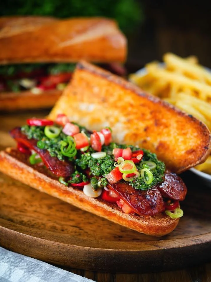 A hot dog with grilled sausage, topped with chopped green onions and relish, served in a toasted bun. In the background, there is a slice of cheese and a side of French fries.