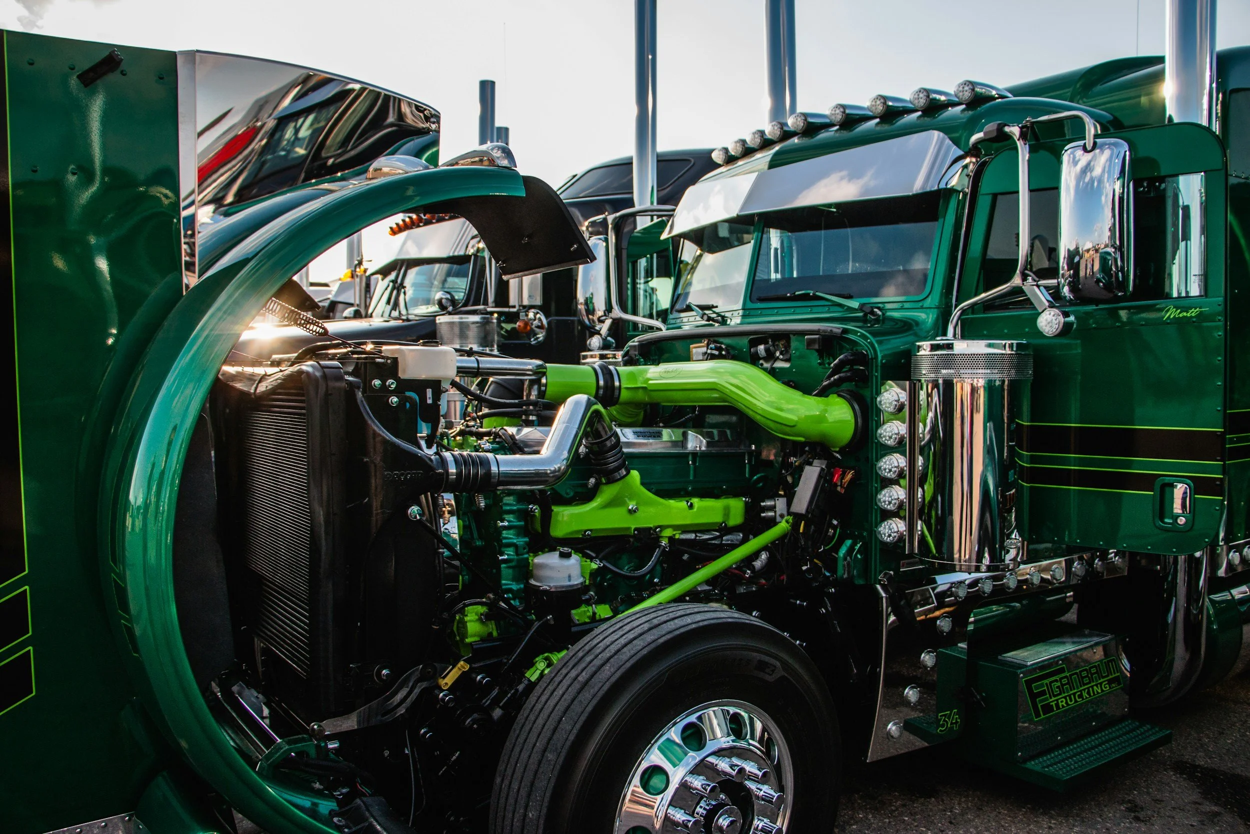 Close-up of a green semi-truck with its hood open, revealing the engine and mechanical components, parked outside under a partly cloudy sky.
