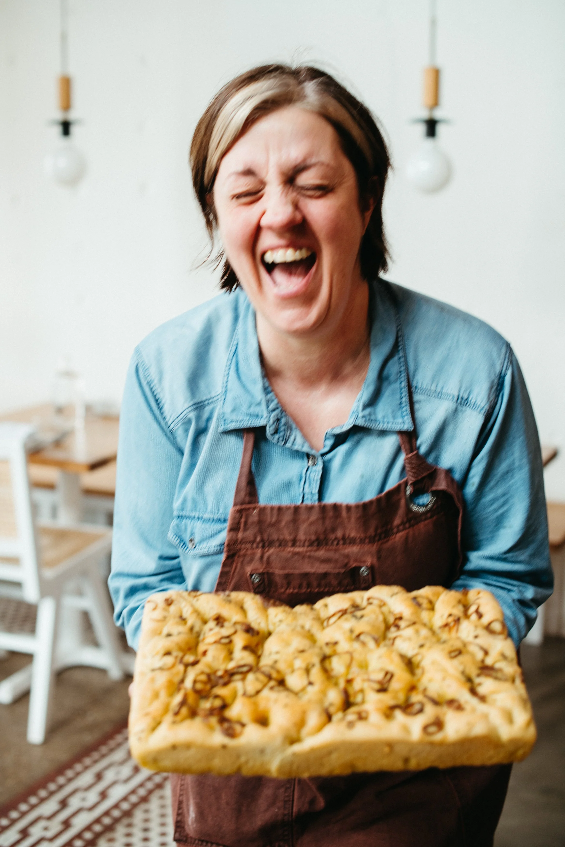 Woman wearing a denim shirt and brown apron holding a tray of freshly baked cookies, smiling with her eyes closed in a cozy kitchen setting.