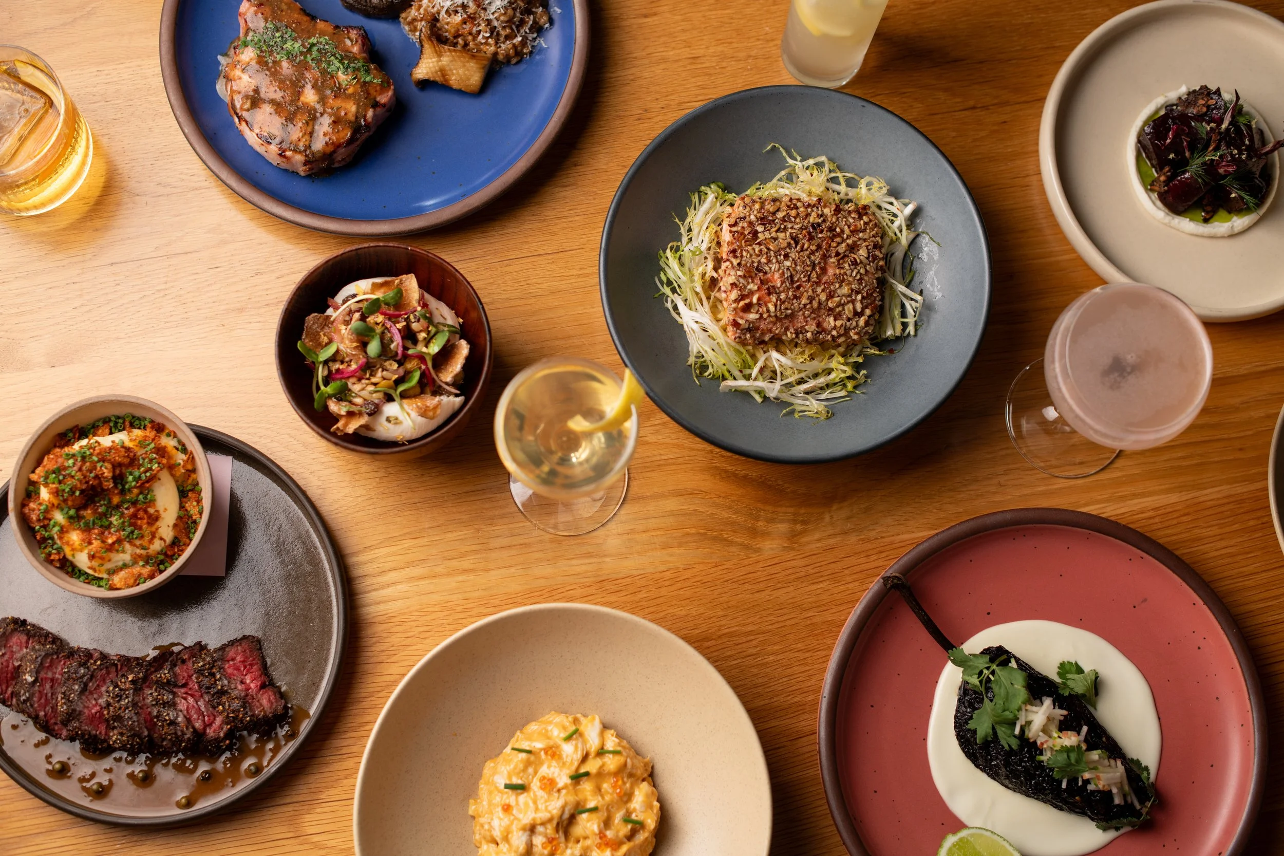 A variety of dishes including salads, grilled meats, and soups on a wooden table, accompanied by lemon slices and beverages.