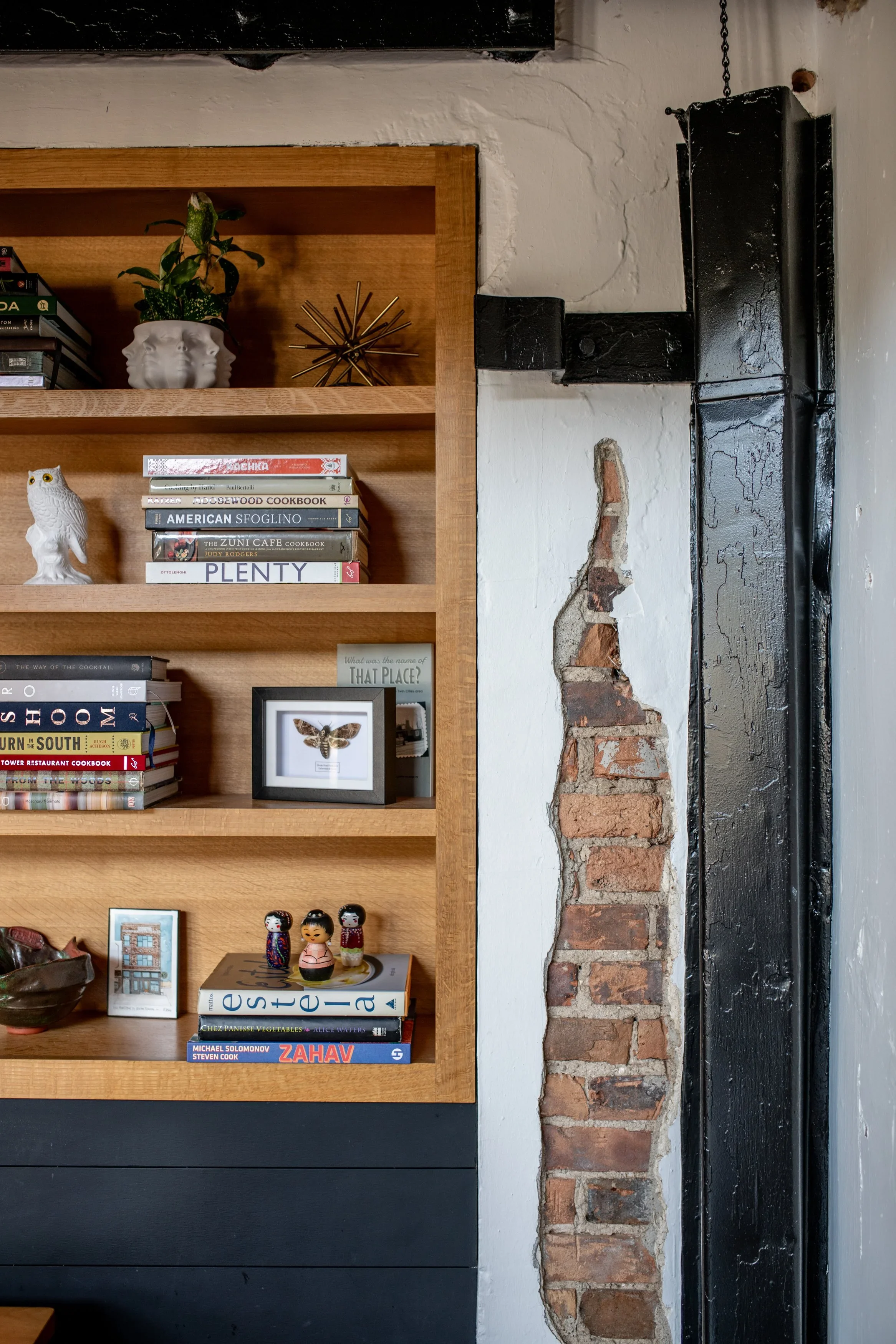 A wooden bookshelf with decorative objects and books against a white textured wall with a black metal structure and exposed brick. Items include a white face-shaped planter with a green plant, a gold starburst sculpture, a white owl figurine, framed art, and various books.