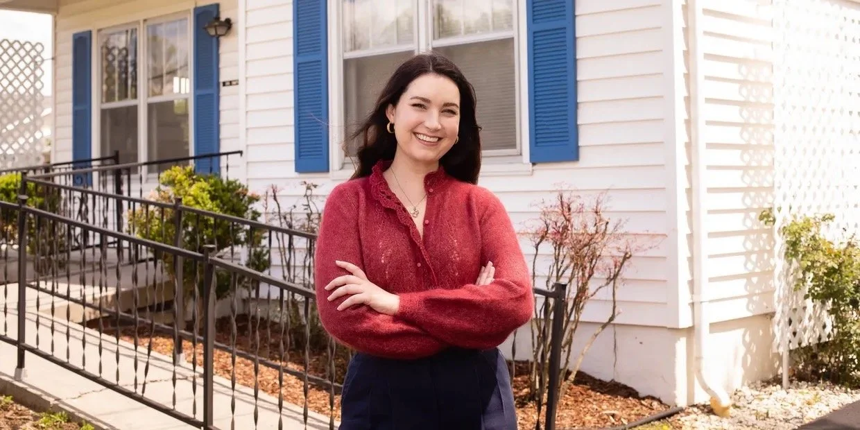 Dr. Kate Palmer standing outside in front of a white house with blue shutters, smiling and crossing her arms
