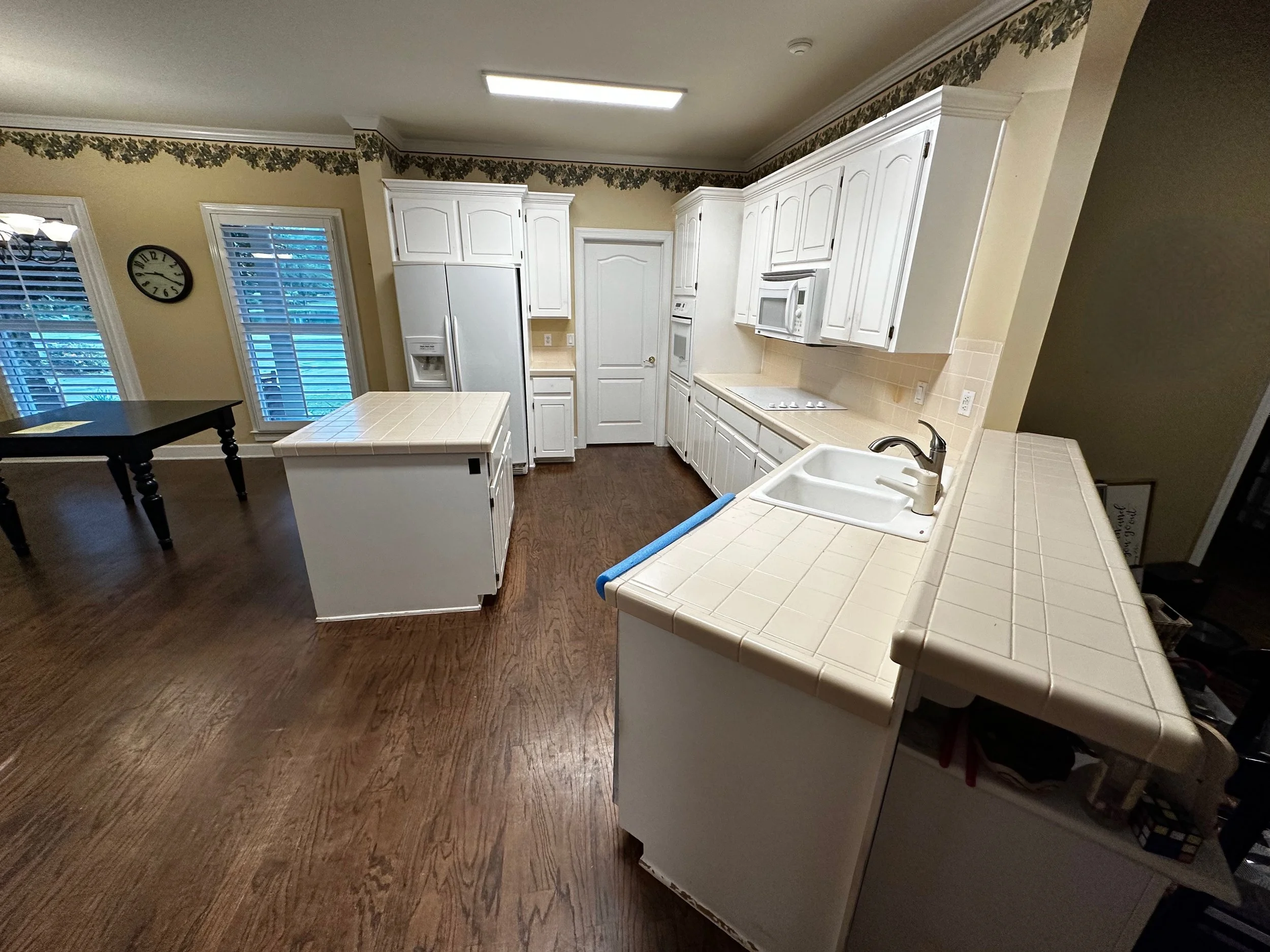 Kitchen with white cabinets, a white refrigerator, a stove, a microwave, and a double sink, with wooden flooring and windows with blinds.