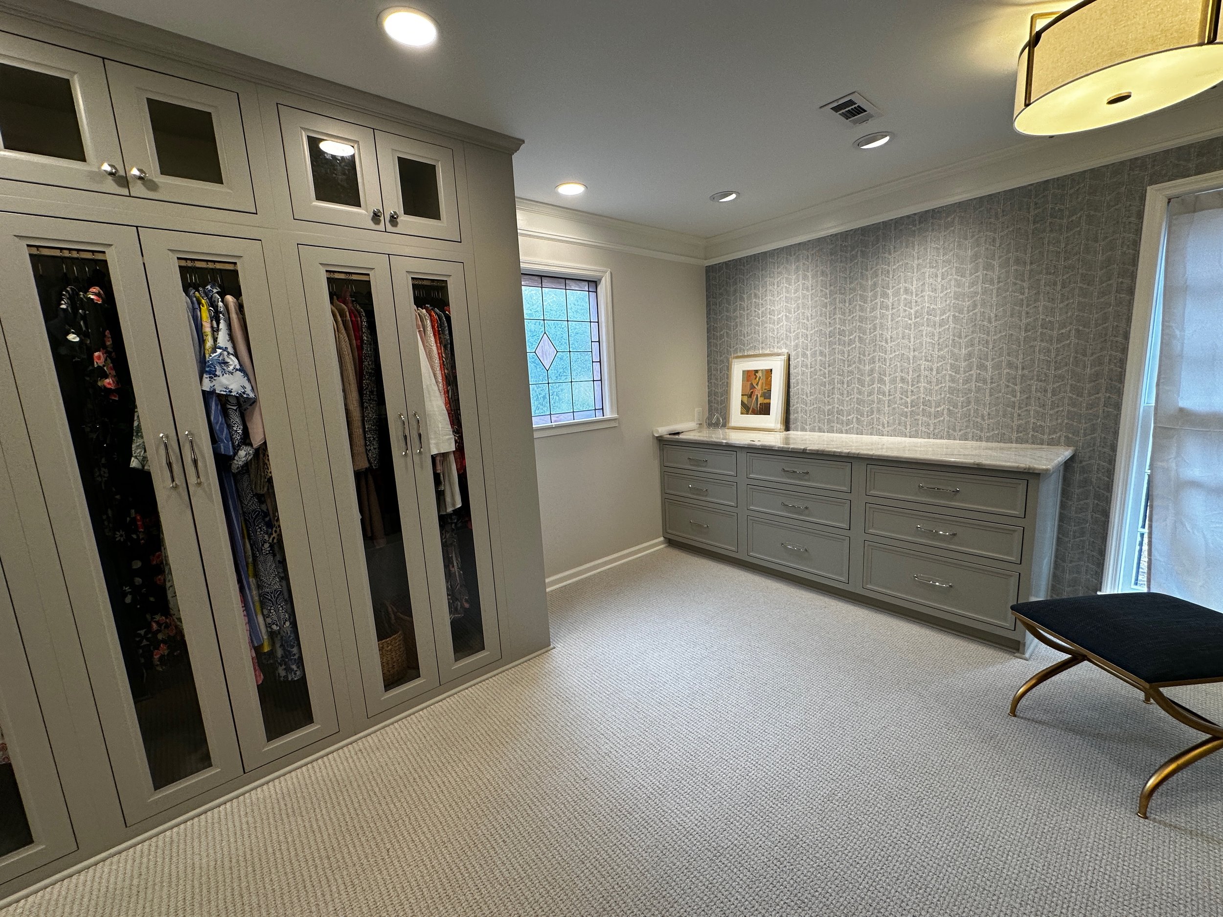 A walk-in closet with closed cabinets with glass panels on the left, filled with hanging clothes. A gray dresser with multiple drawers and a marble top is against a textured patterned gray wallpapered wall. A framed artwork and a small white object a