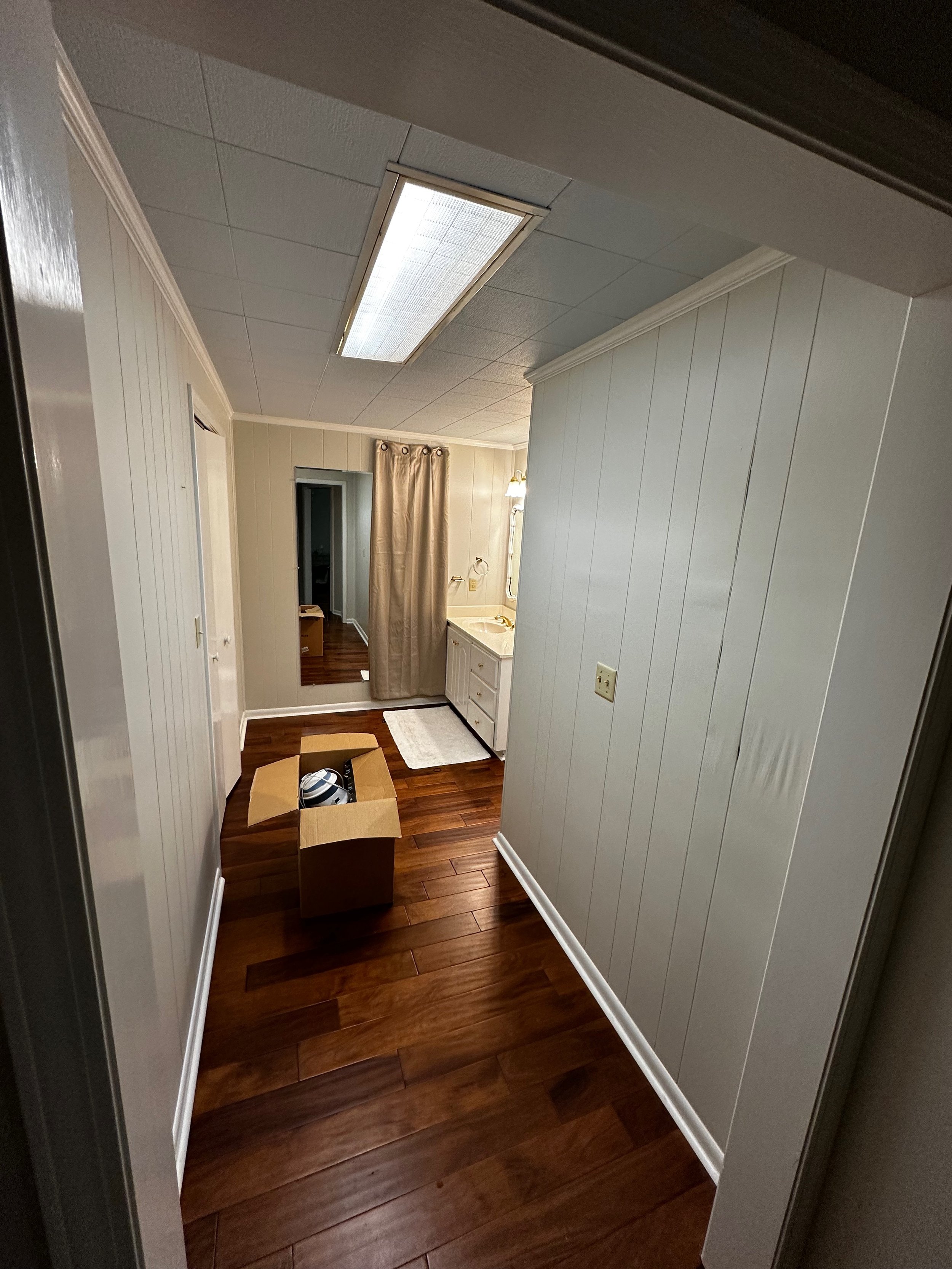 View of a hallway or small room with wood flooring, cream-colored paneled walls, a ceiling with a fluorescent light, and a doorway with a curtain. There is a cardboard box on the floor with items inside, a small white rug near a kitchenette with a si