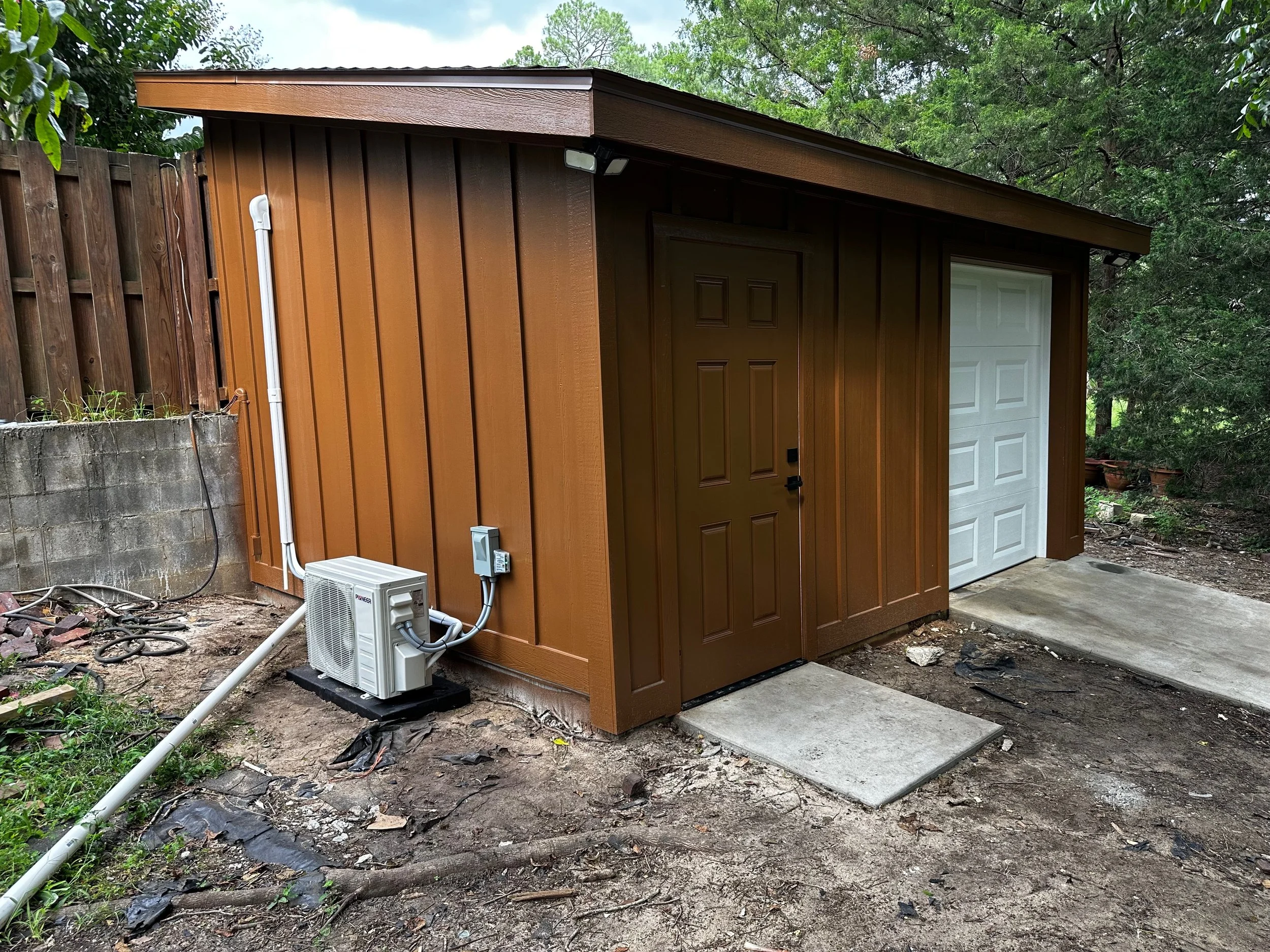 A small brown shed with a white garage door and a brown door, surrounded by trees and a dirt ground with a concrete slab in front of each door. An air conditioning unit is installed outside the shed.
