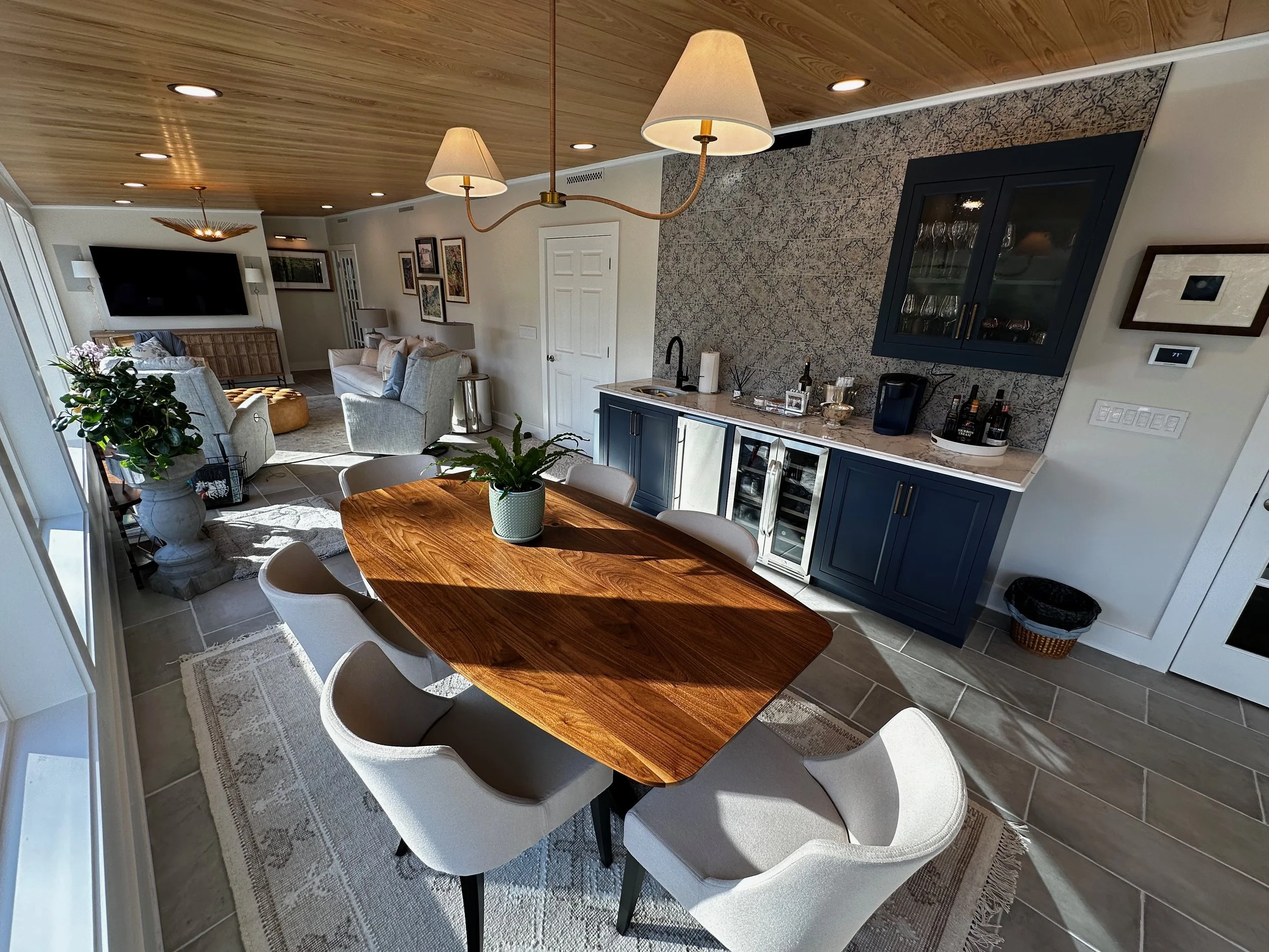 Open-concept living and dining area with a wooden dining table, six upholstered chairs, a built-in blue and white bar area, a beige rug, a large window, and a cozy seating space with armchairs and a television.