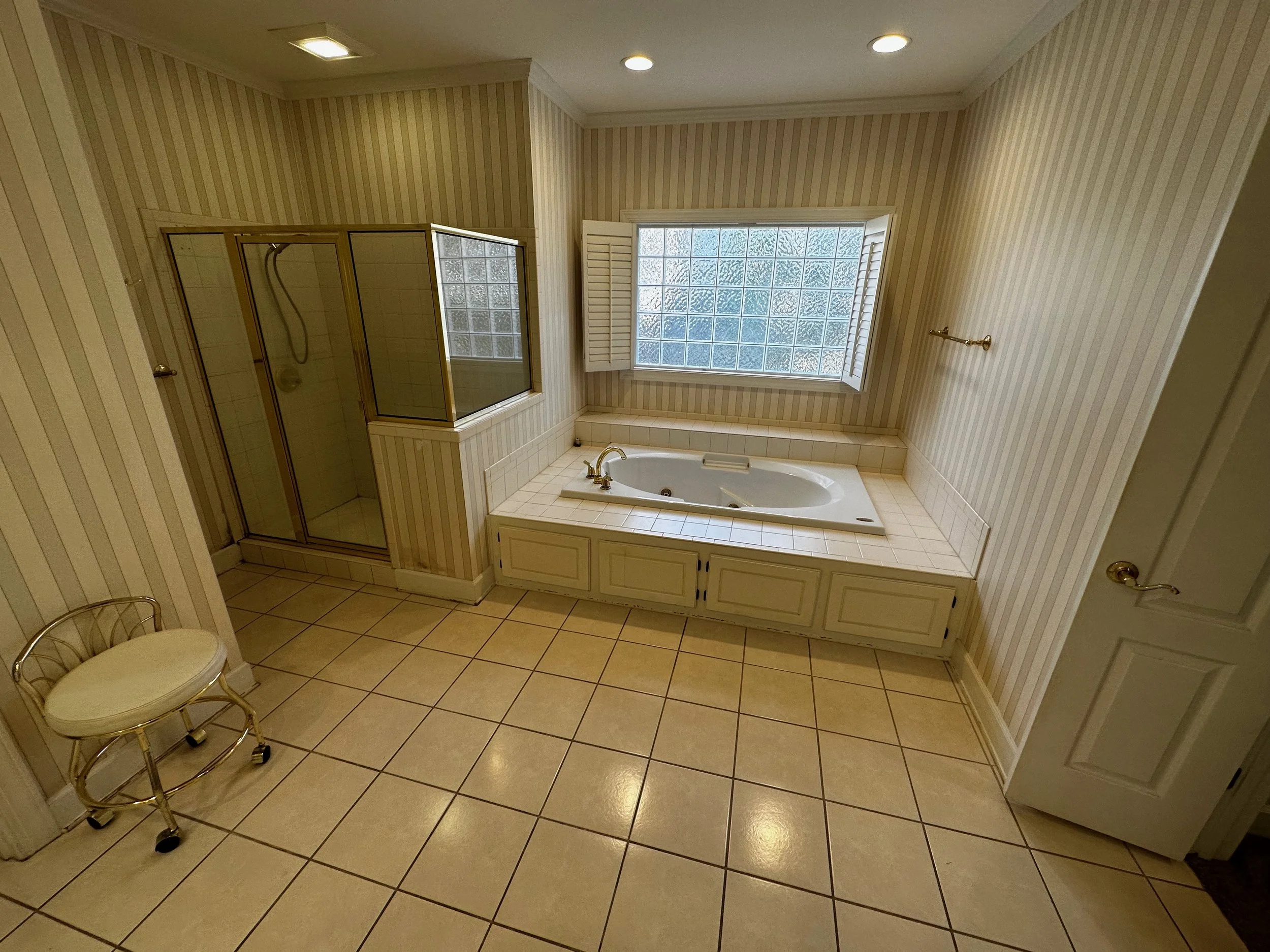 Bathroom with a walk-in shower and a bathtub near a large frosted glass window, beige tiled floor, striped wallpaper, a small chair with a white cushion, and a towel rack in the background.