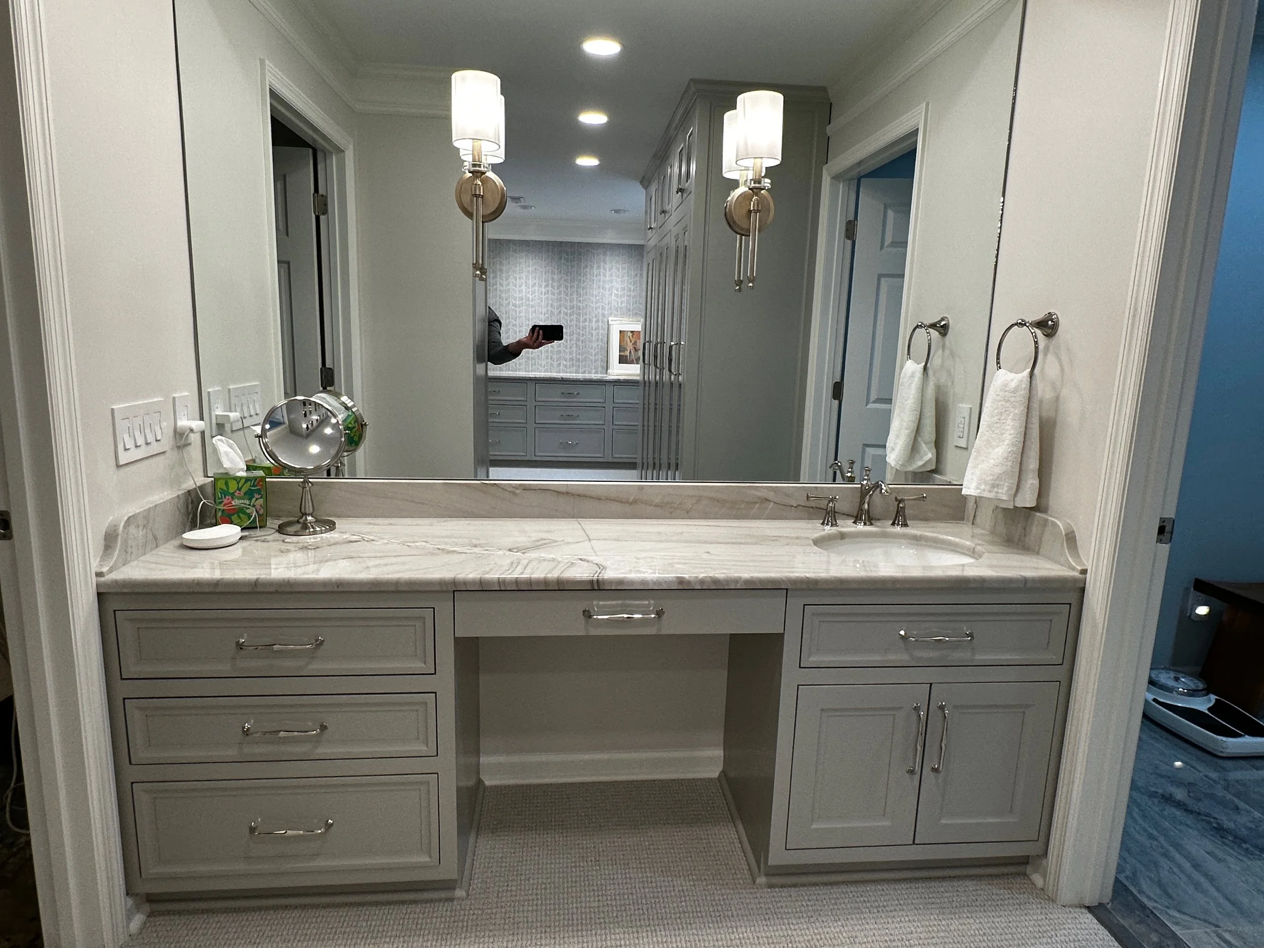Bathroom vanity with marble countertop, oval sink, silver faucet, three drawers and a cabinet, two wall-mounted towel rings with white towels, decorative light fixtures, mirror, and tissue box with a tissue, in a modern bathroom.
