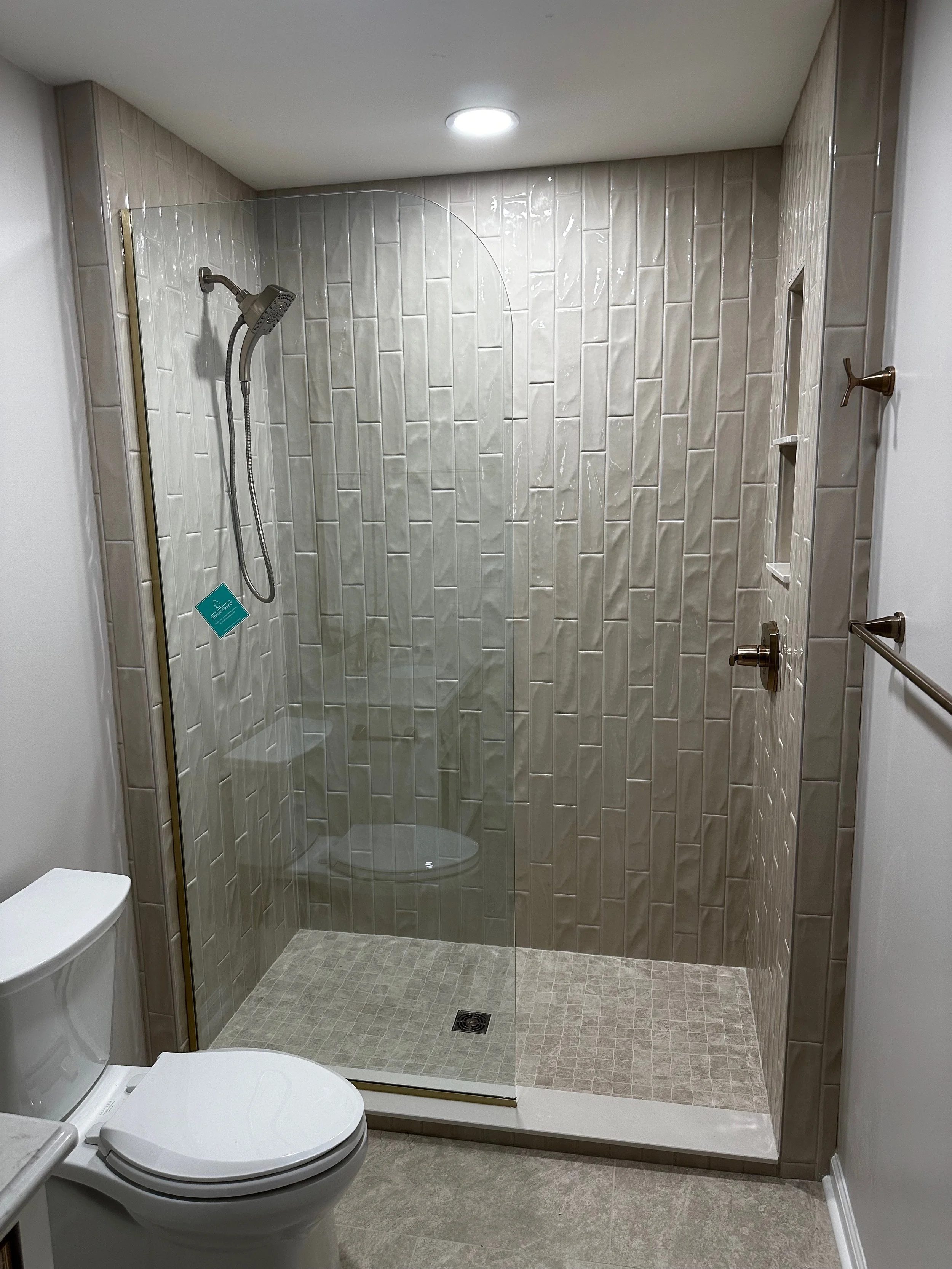 Bathroom with a walk-in shower that has beige tiles, a glass door, a handheld showerhead, built-in shelves, a towel holder, and a white toilet.