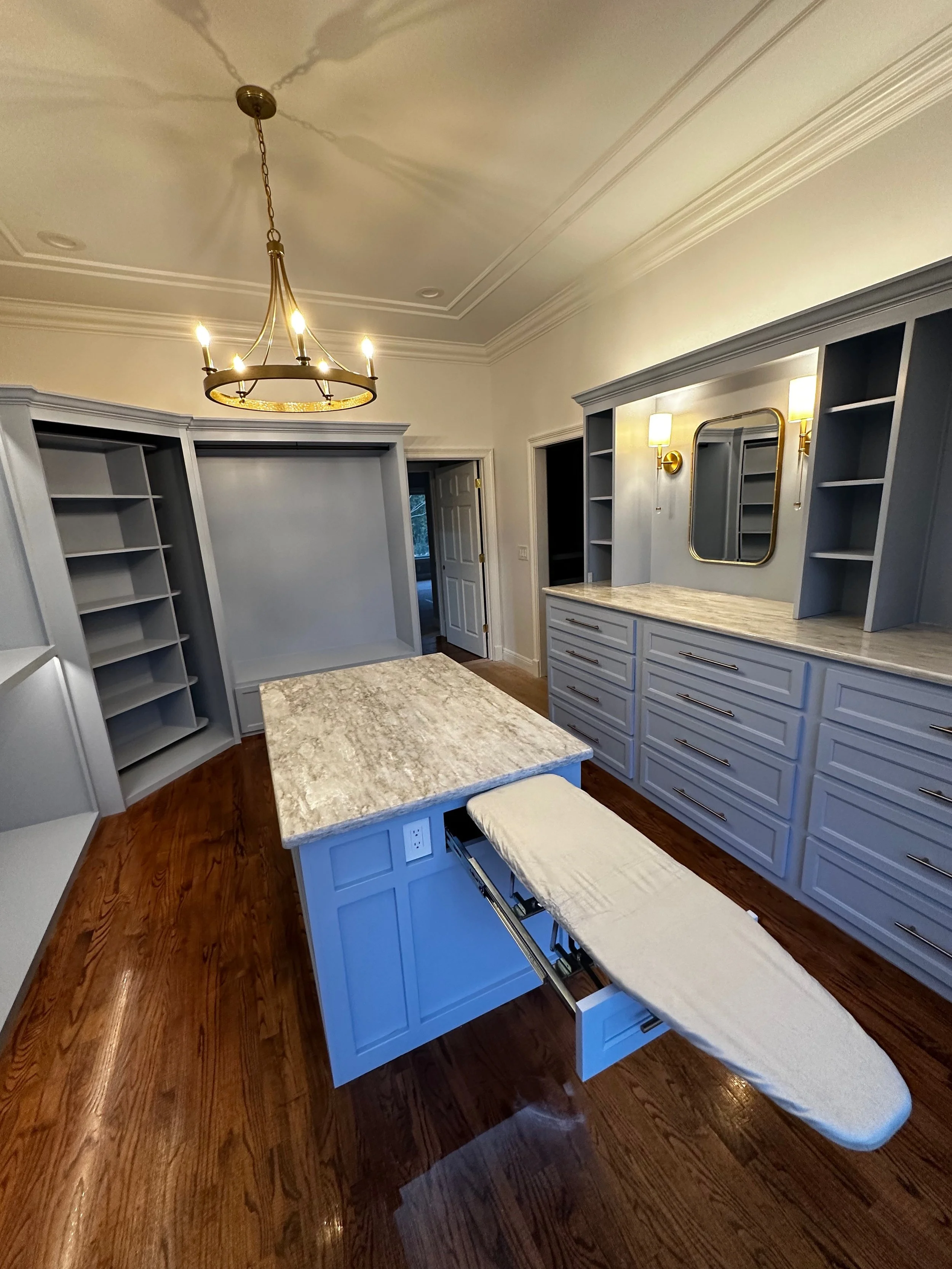 A walk-in closet with built-in shelving, drawers, and a central island with a marble top. There is a wall mirror, two wall sconces, a chandelier, and hardwood floors.