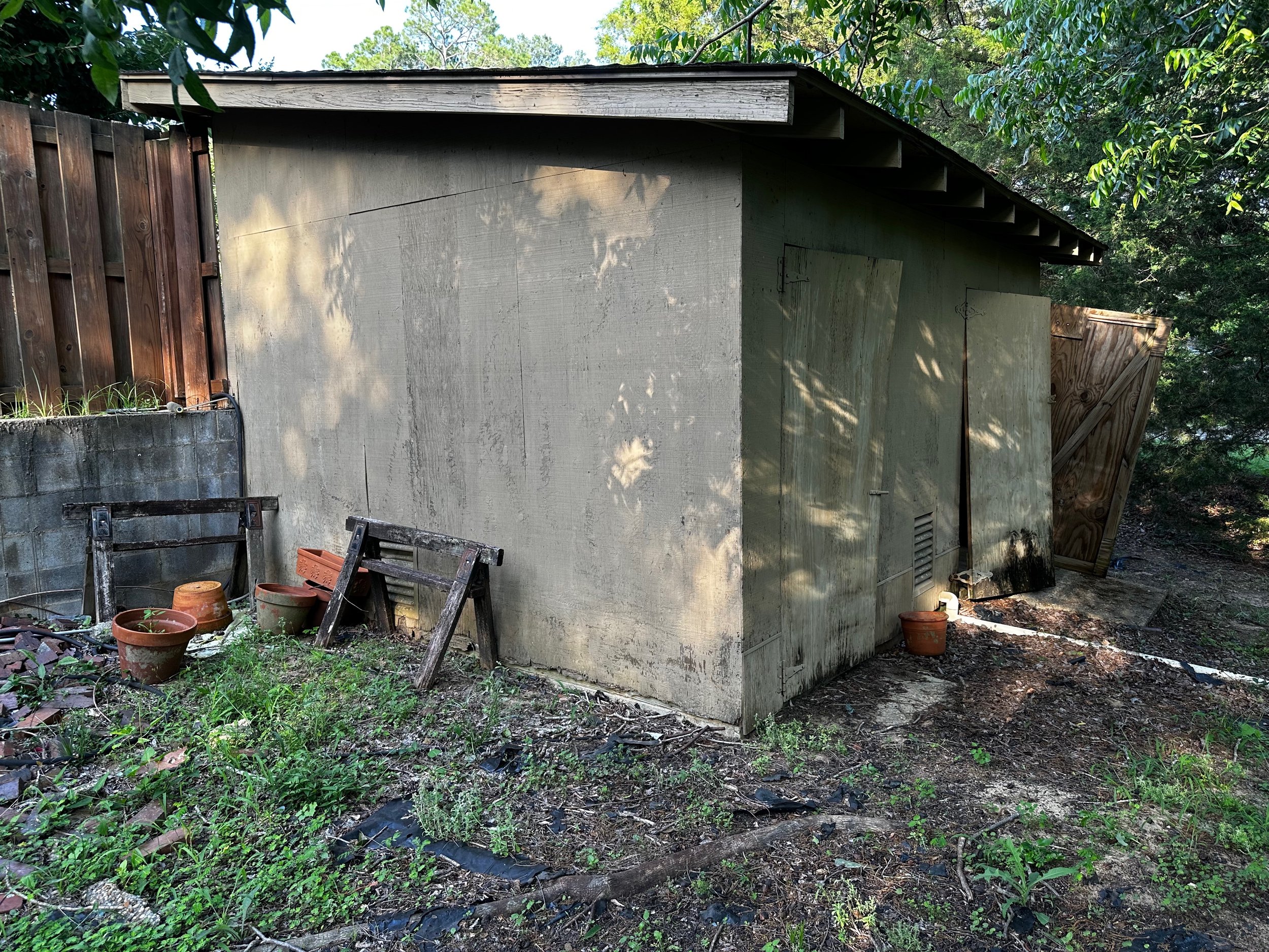 A small, weathered shed on a patch of dirt and grass, surrounded by trees and wooden fencing, with gardening pots and wooden pallets leaning against the shed.