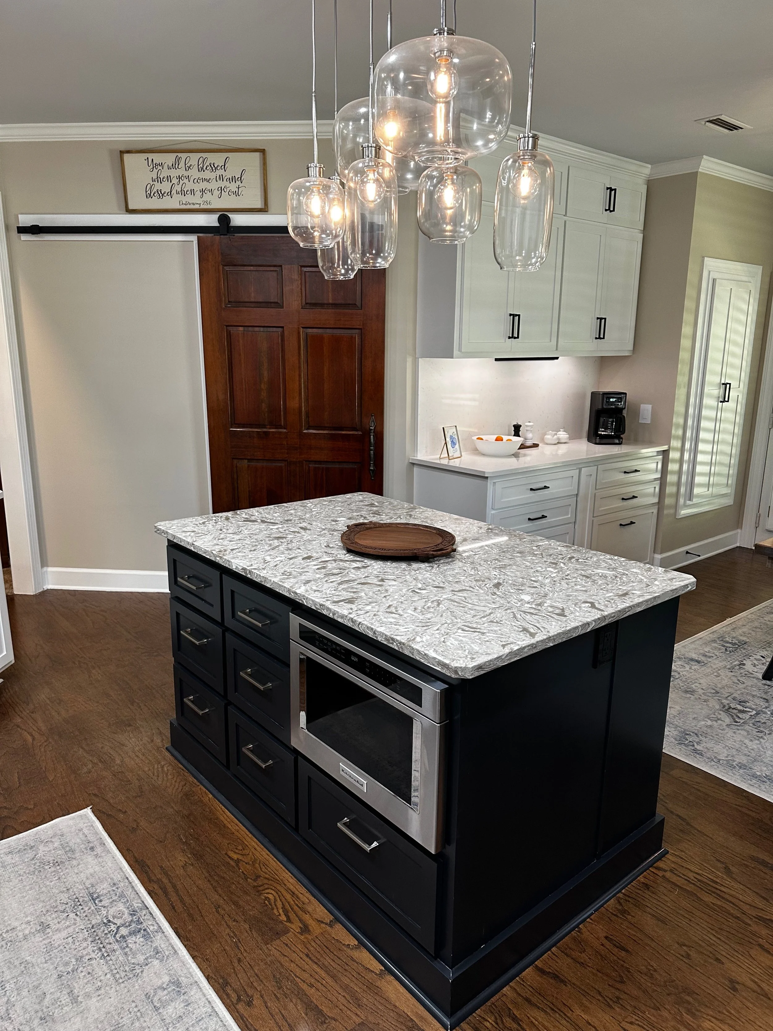 Modern kitchen with a black island featuring a marble countertop, overhanging glass pendant lights, white cabinets, and a wooden door.