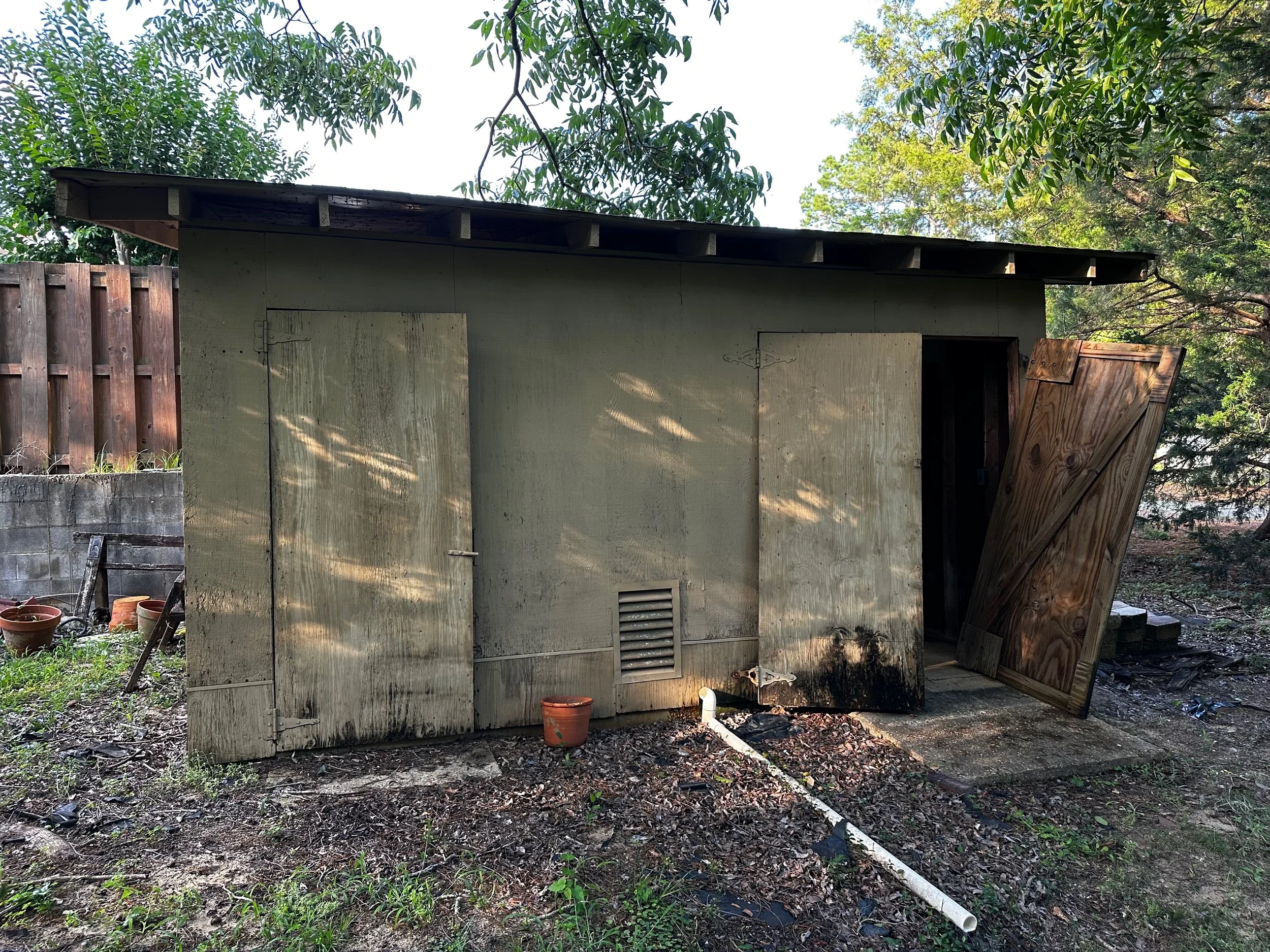 A small, weathered shed with two closed and one partly open door, one of which is made of unpainted wood. The shed has a vent near the bottom, along with some black staining on the lower part of the walls. Surrounding the shed are trees with green le
