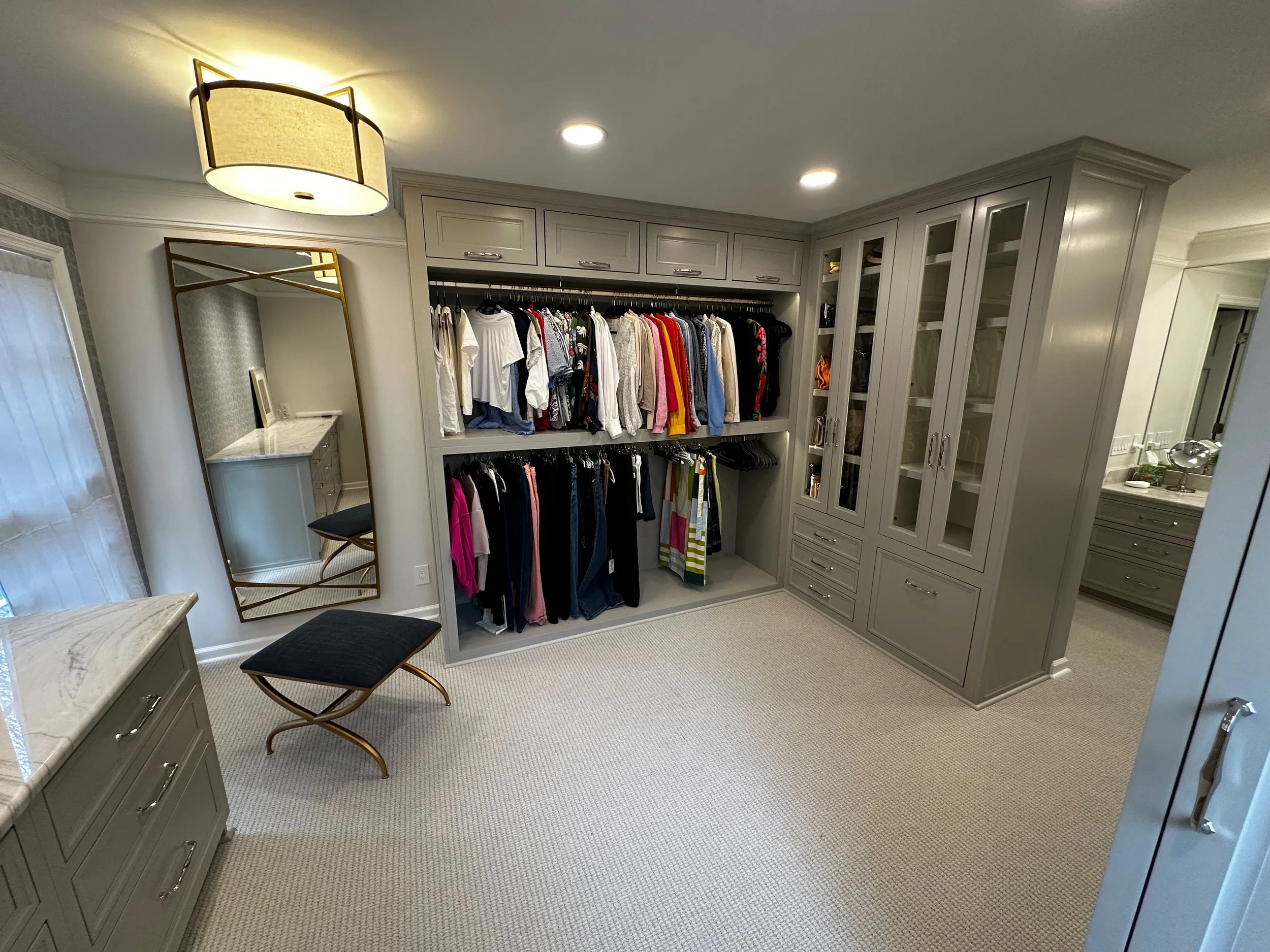 A walk-in closet with built-in gray cabinetry and an open closet section with hanging clothes. There is a mirror on the wall, a bench with a dark cushion, and a marble-topped dresser. The room has beige carpeting and a ceiling light fixture.