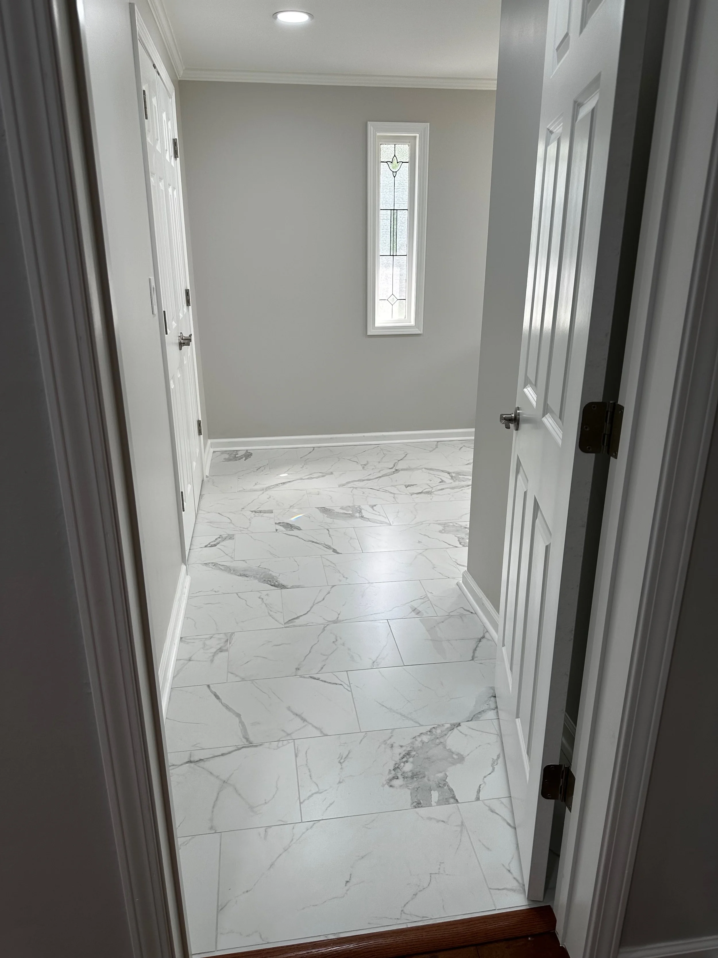 Empty room with white marble floor, light gray painted walls, a tall, narrow window with stained glass, and a closed door on the right side