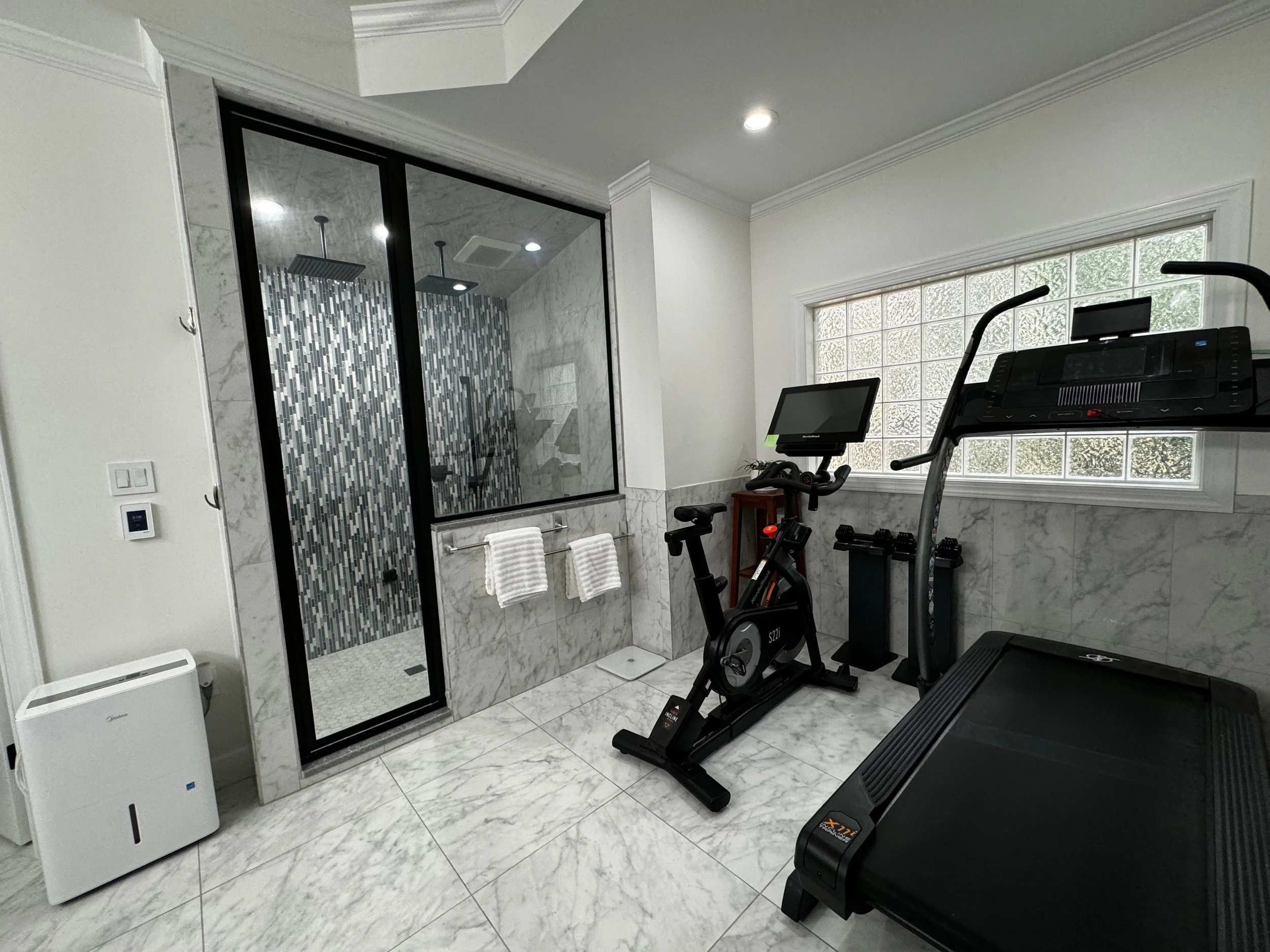 Home gym area with treadmill, stationary bike, and an adjacent walk-in shower enclosed with glass, featuring marble walls and gray tile flooring.