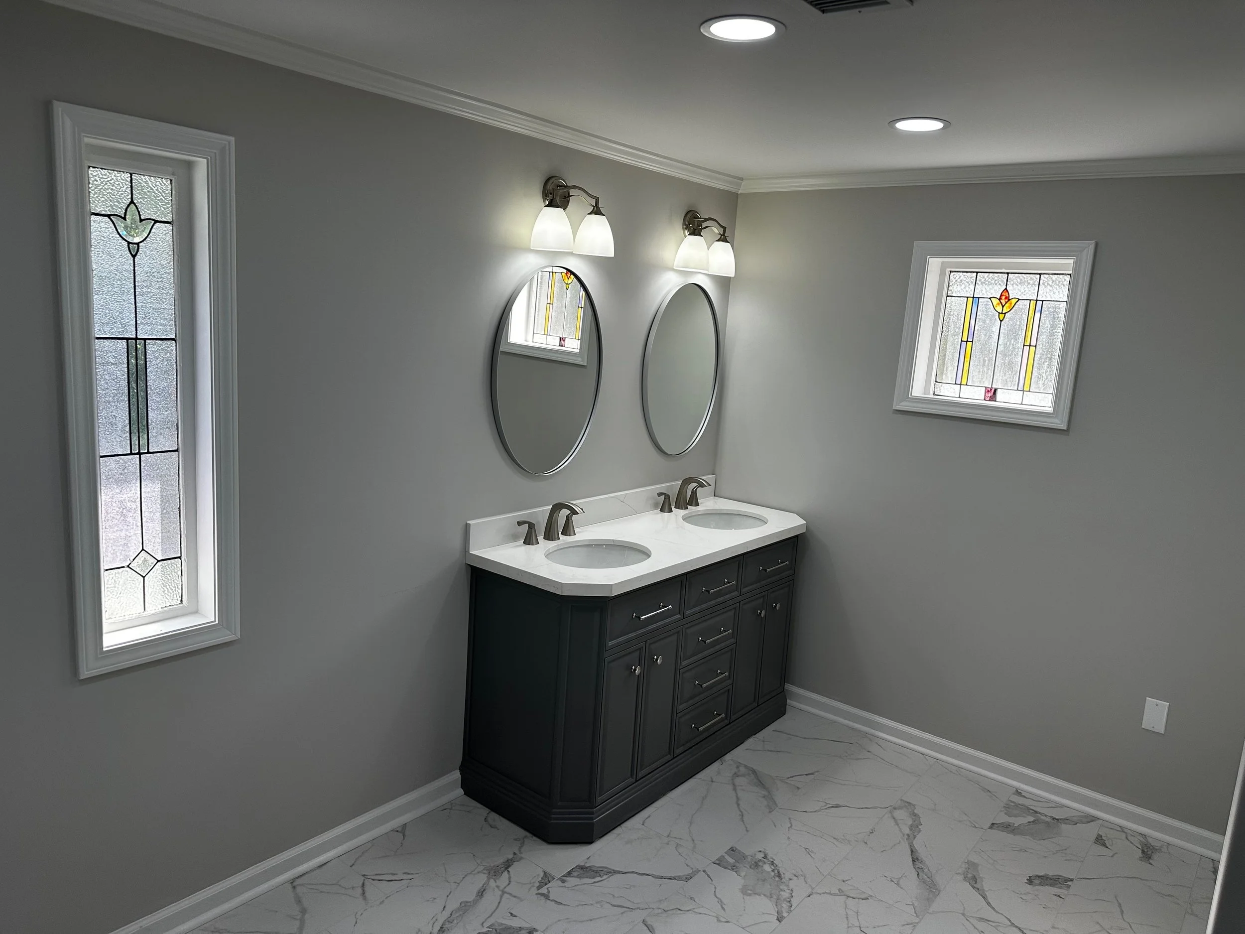 A modern bathroom with a dark vanity, two oval mirrors, and stained glass windows, illuminated by ceiling and wall lights.