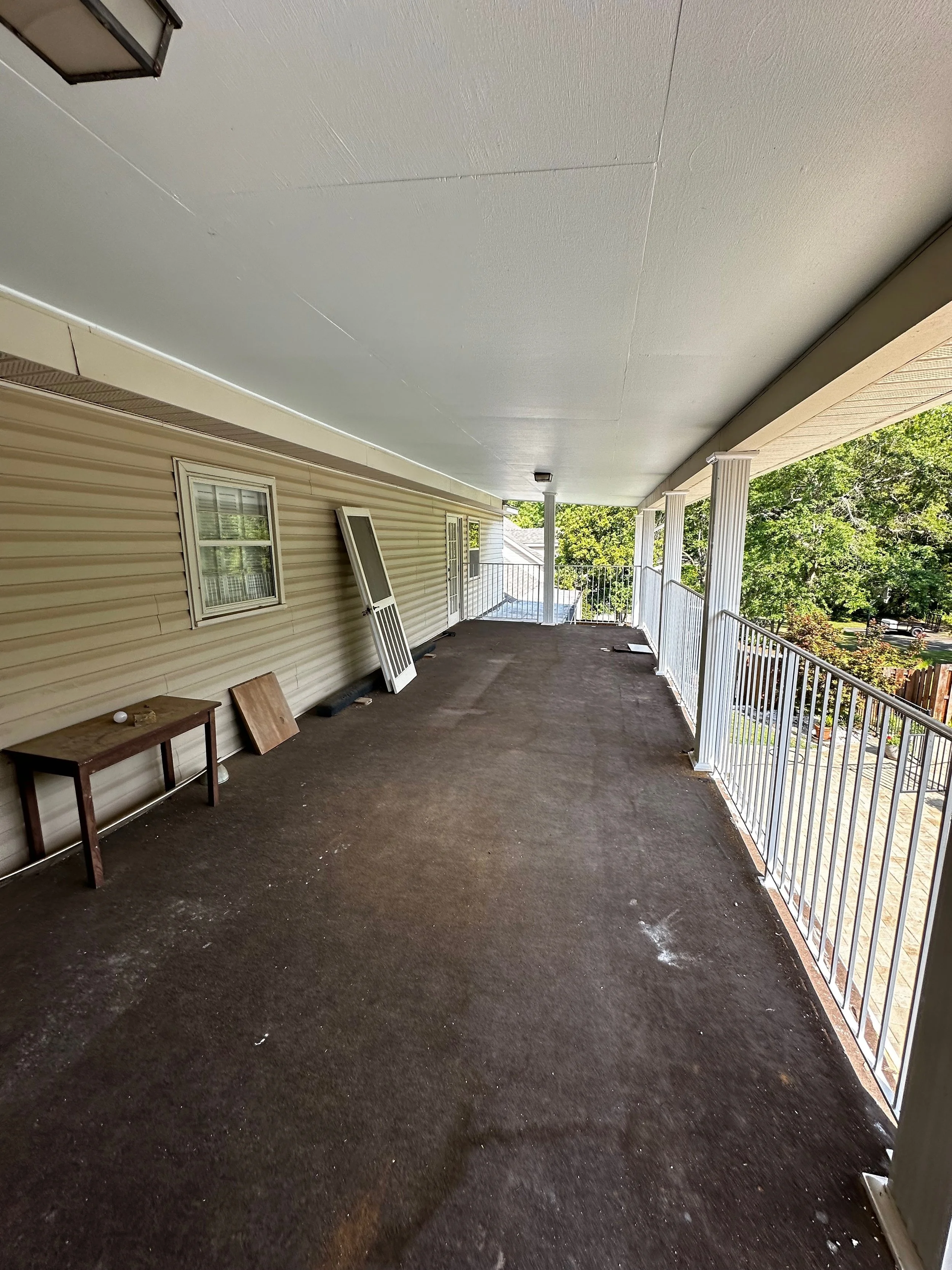 Long outdoor porch with dark flooring, white railing, and beige siding, with tools and window screens leaning against the wall.