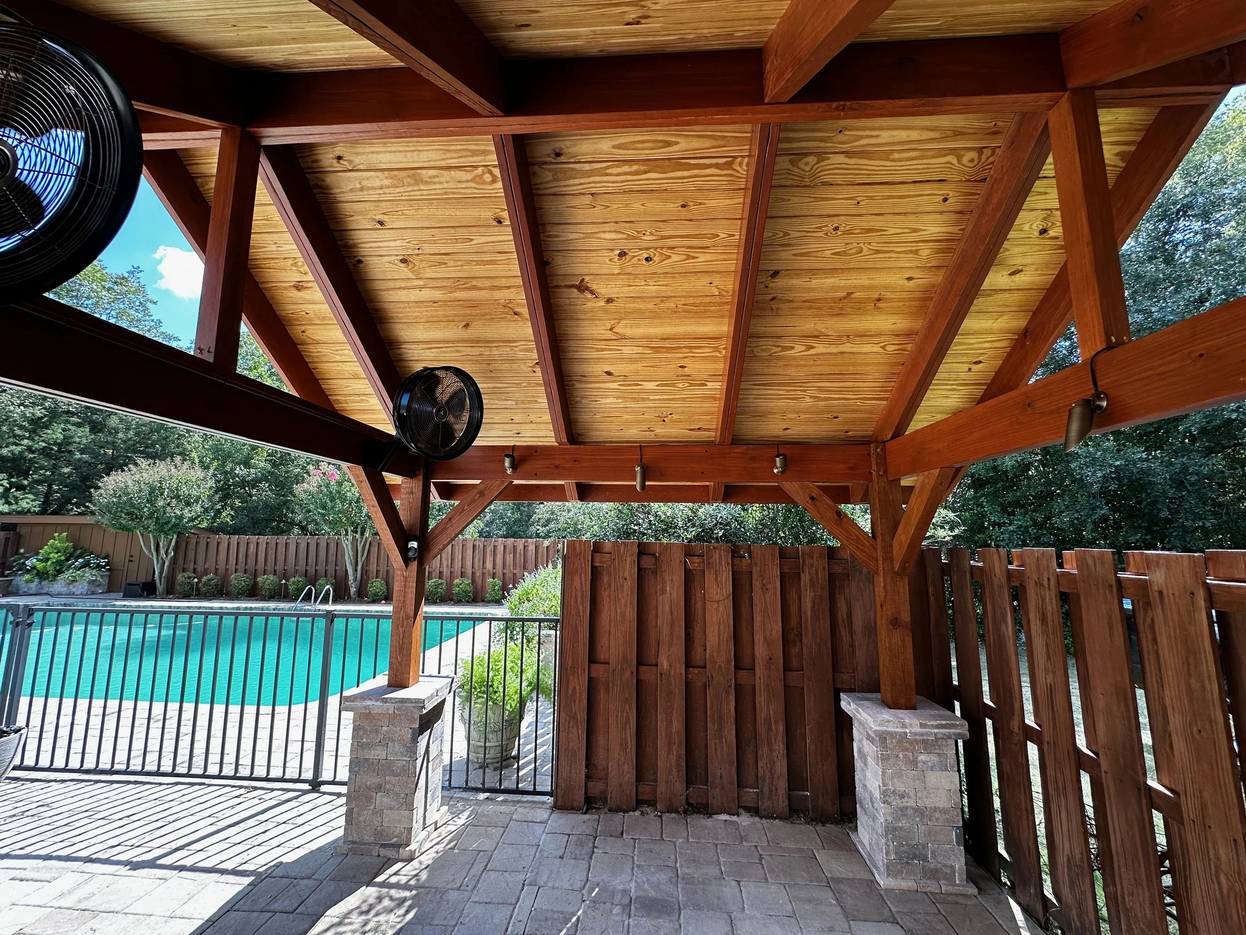 Under a wooden pergola overlooking a backyard swimming pool with a black metal fence, brick columns, and trees in the background.